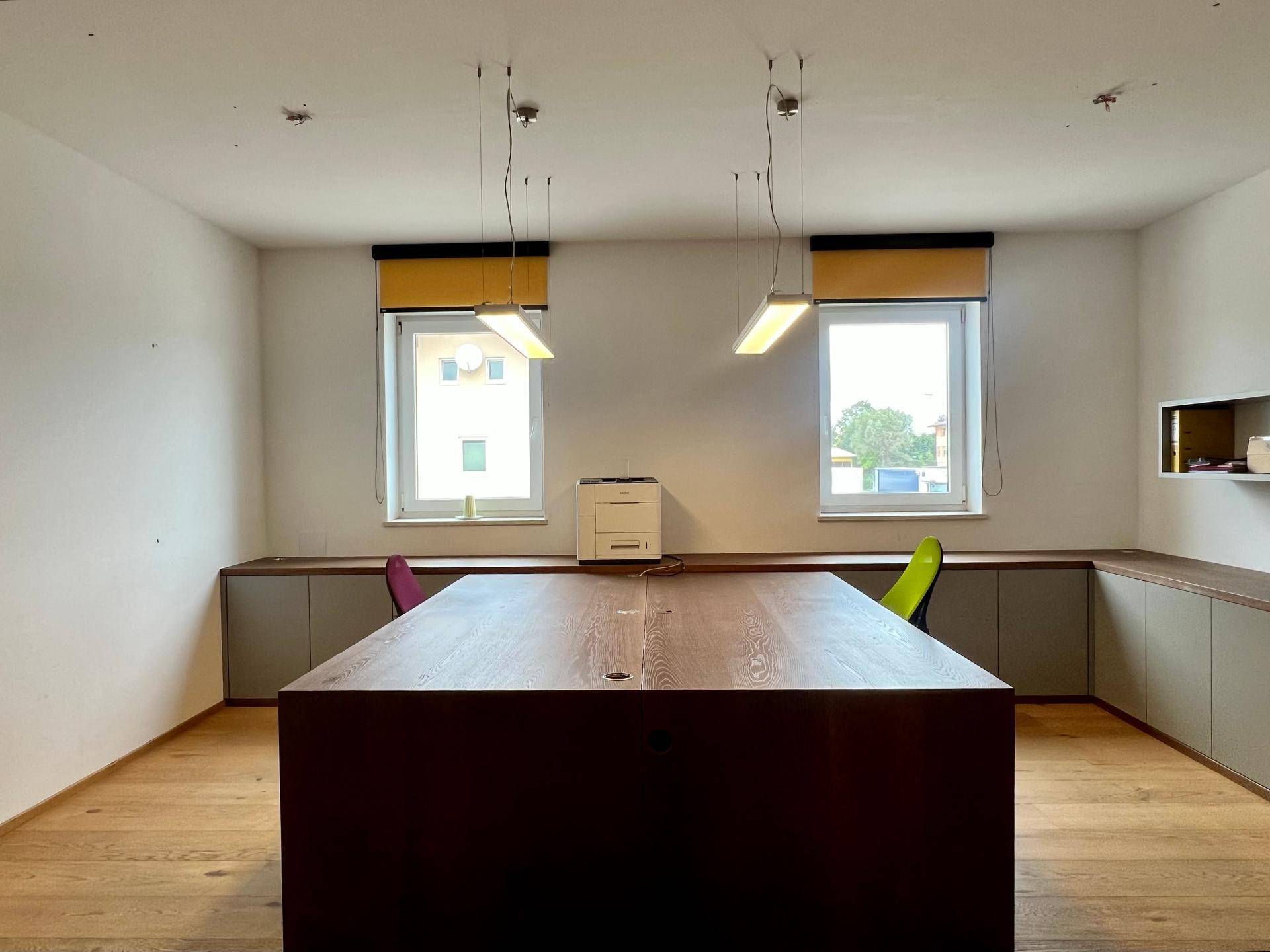 Heller Büroraum mit großem Holztisch, zwei Fenstern und integrierten Sideboards für effizientes Arbeiten.