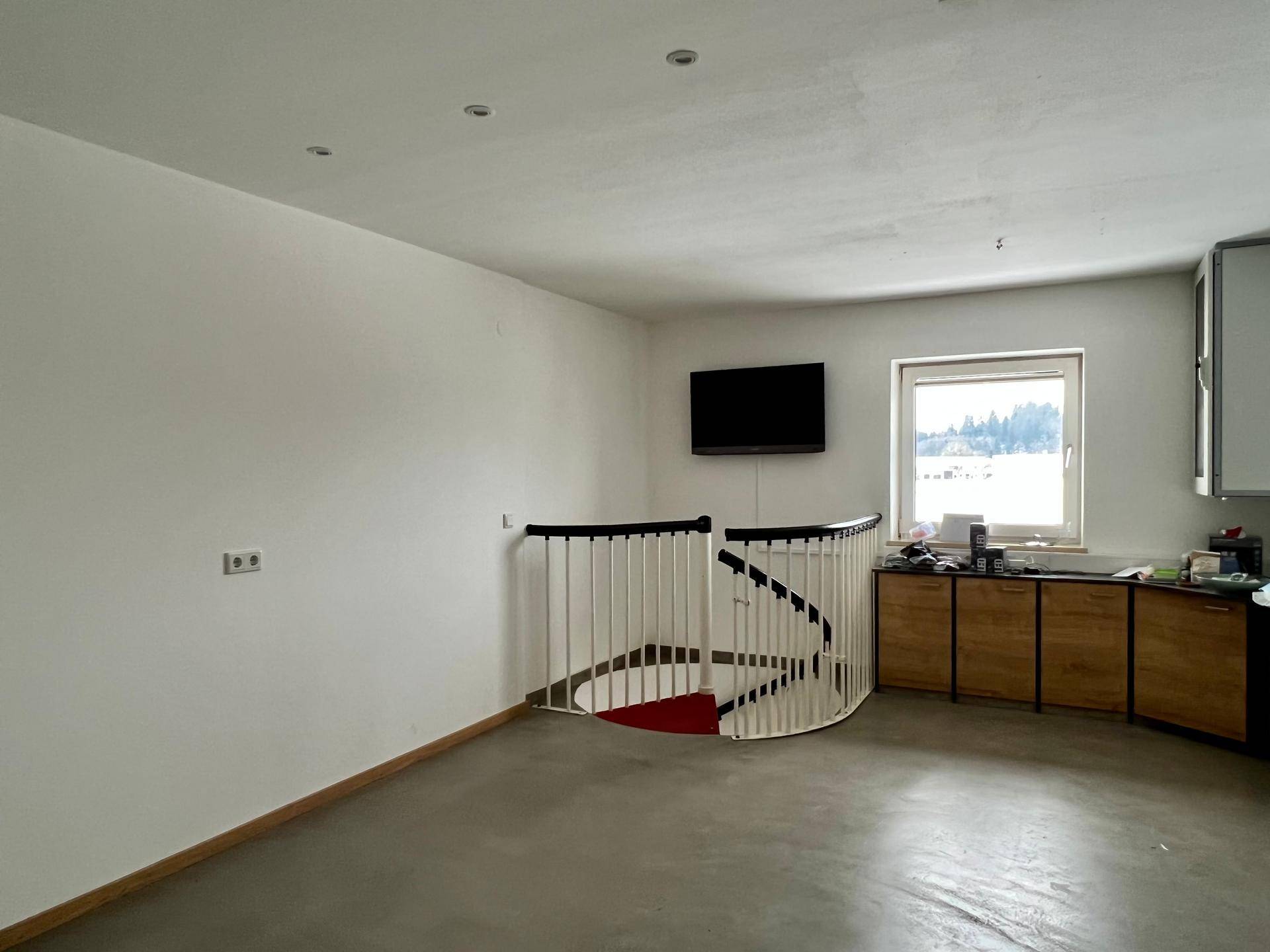 Geräumiger Raum mit einer Wendeltreppe, einem Wandfernseher und einer Küchenzeile im Hintergrund.
