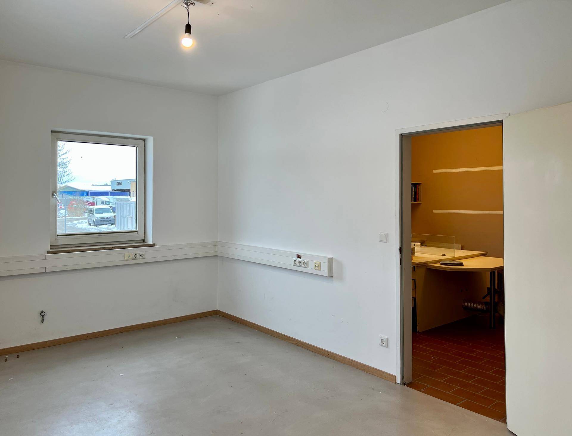 Heller Büroraum mit einem Fenster, weißen Wänden und einer offenen Tür zu einem weiteren Raum.