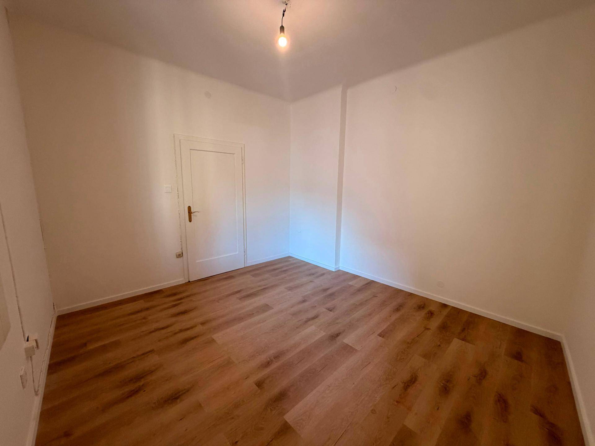 Helles, leeres Zimmer mit Laminatboden und weißer Tür, bereit für individuelle Gestaltungsmöglichkeiten.