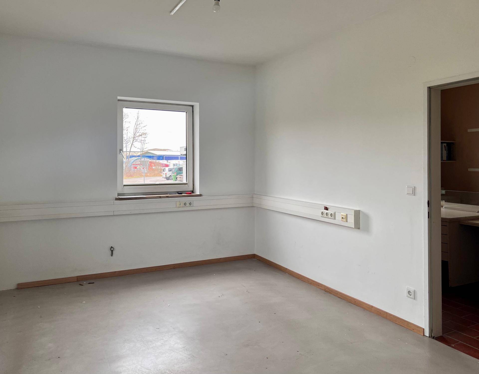 Leerer Büroraum mit einem Fenster, weißen Wänden und einer Tür zu einem angrenzenden Bereich.