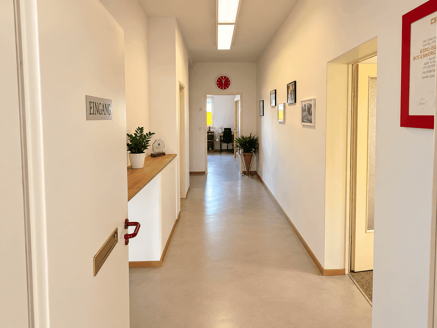 Langer, heller Flur mit weiß gestrichenen Wänden, Pflanzen und Bildern, der zu weiteren Büroräumen führt.