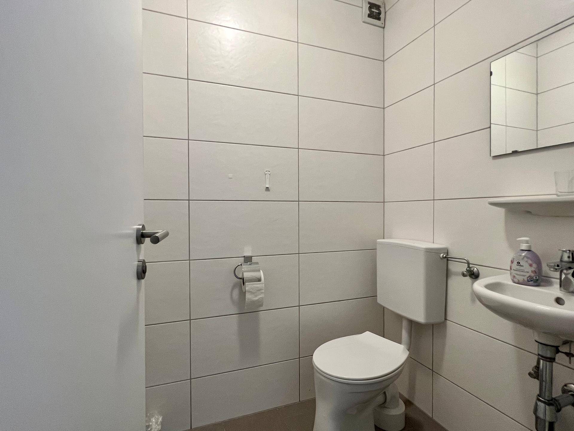 Einfache Gästetoilette mit weißen Fliesen, wandmontierter Toilette und kleinem Waschbecken.