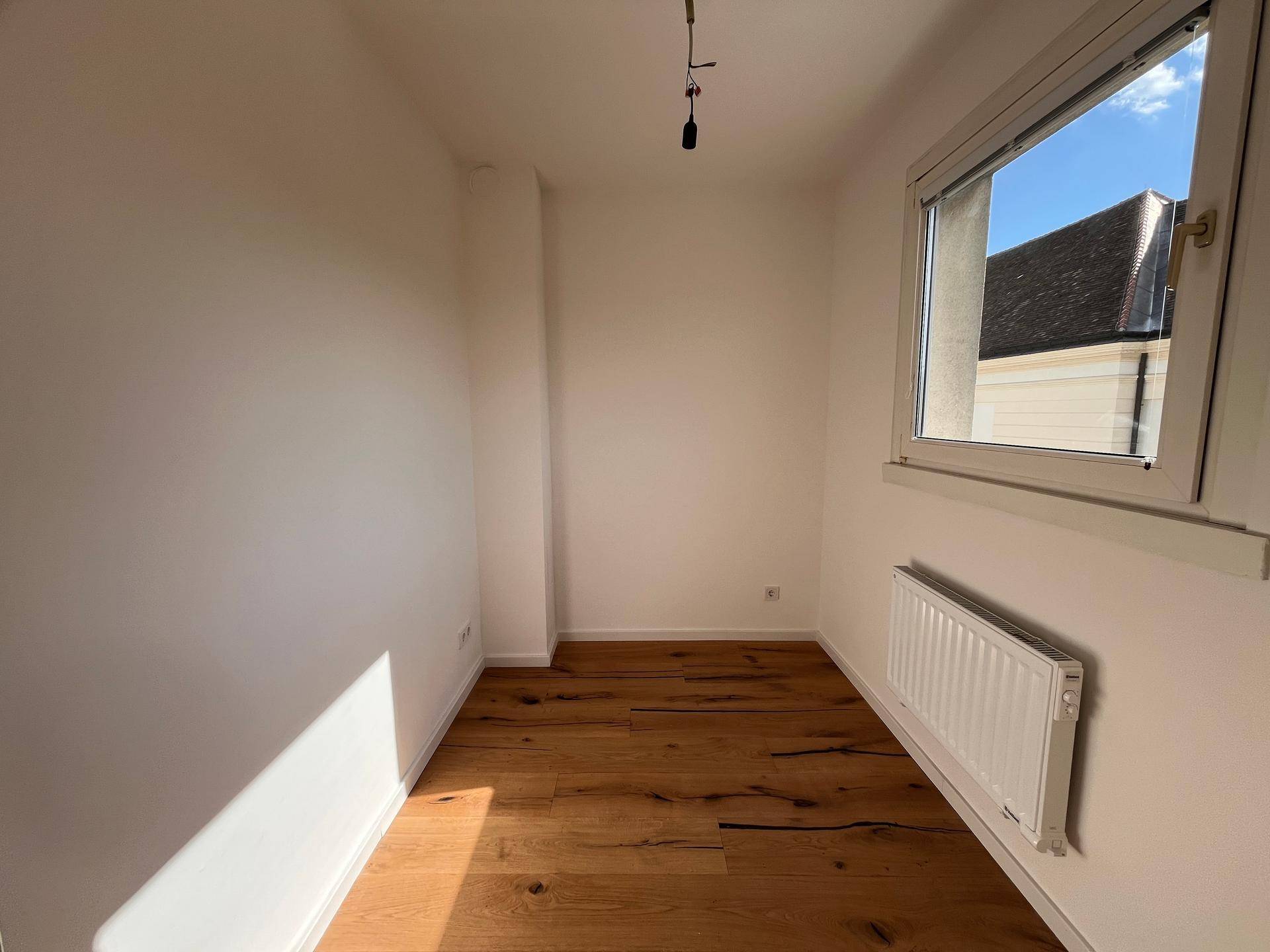 Kleines, helles Zimmer mit Holzboden und Fenster, geeignet als Schlafzimmer oder Arbeitsbereich.