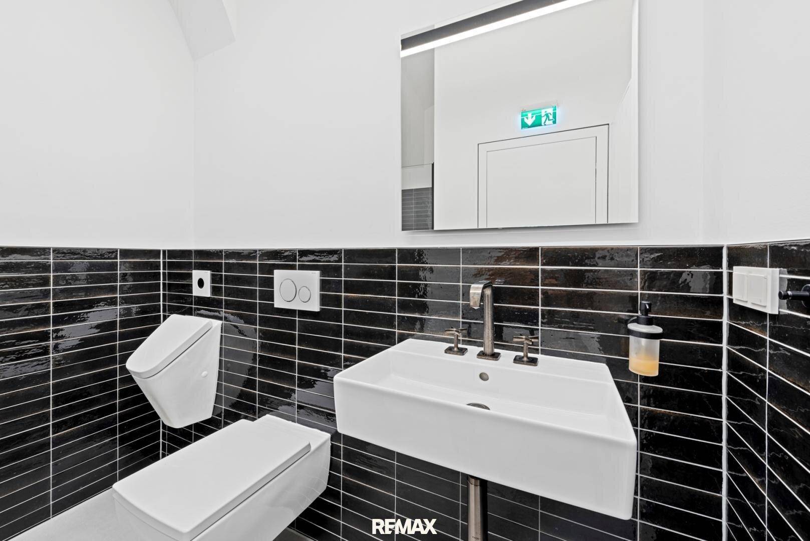 Modernes Badezimmer mit schwarzen Fliesen, weißem Waschbecken und wandhängender Toilette in minimalistischem Design.
