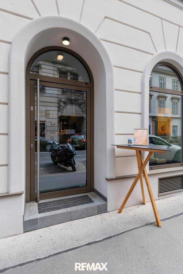 Eingangsbereich eines Gebäudes mit eleganter Bogen-Glastür und einem kleinen Stehtisch davor.