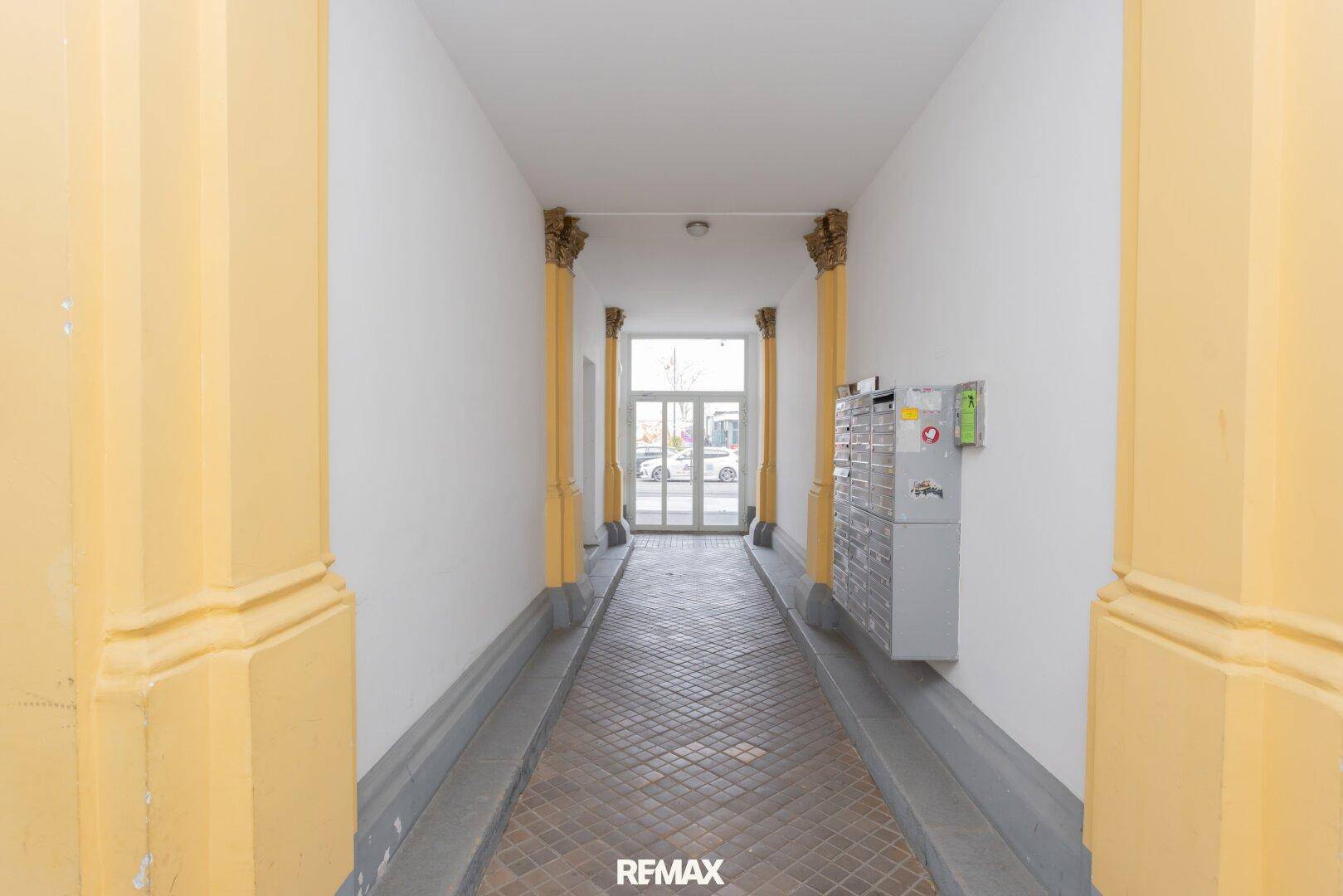 Langer Eingangsbereich mit gelben Säulen, Fliesenboden und Briefkästen an der Wand.