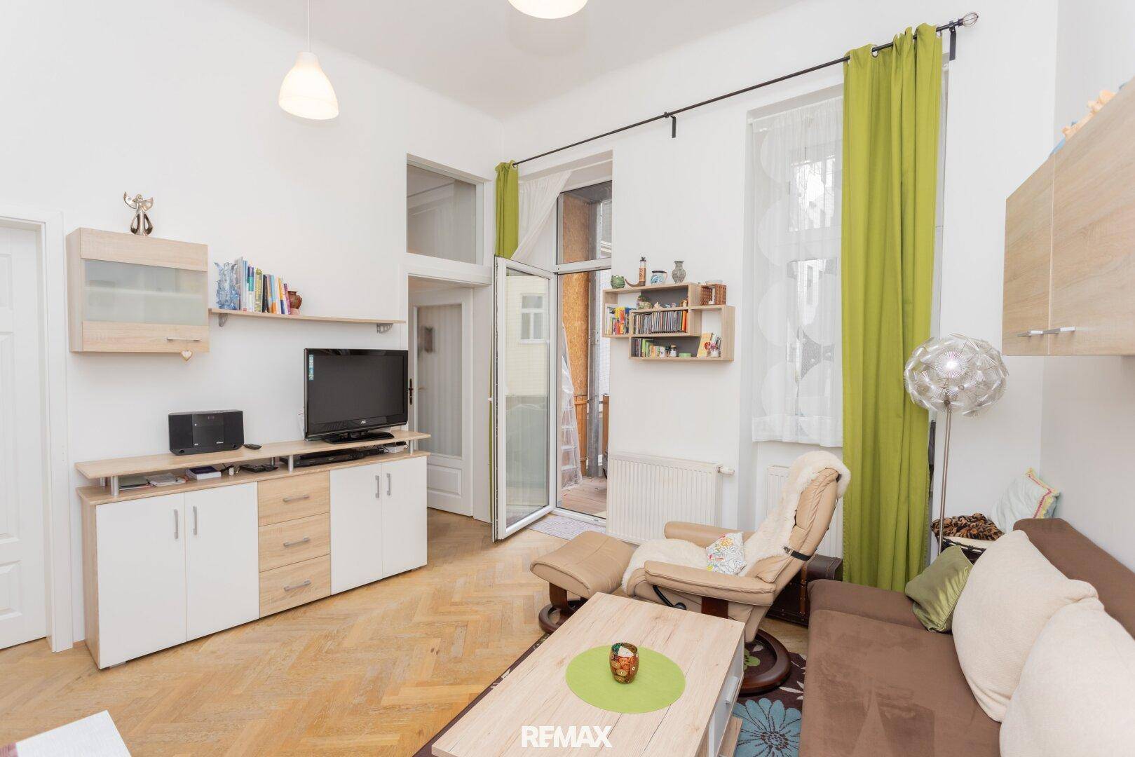 Gemütliches Wohnzimmer mit Fischgrätparkett, Balkonzugang und grünen Vorhängen.
