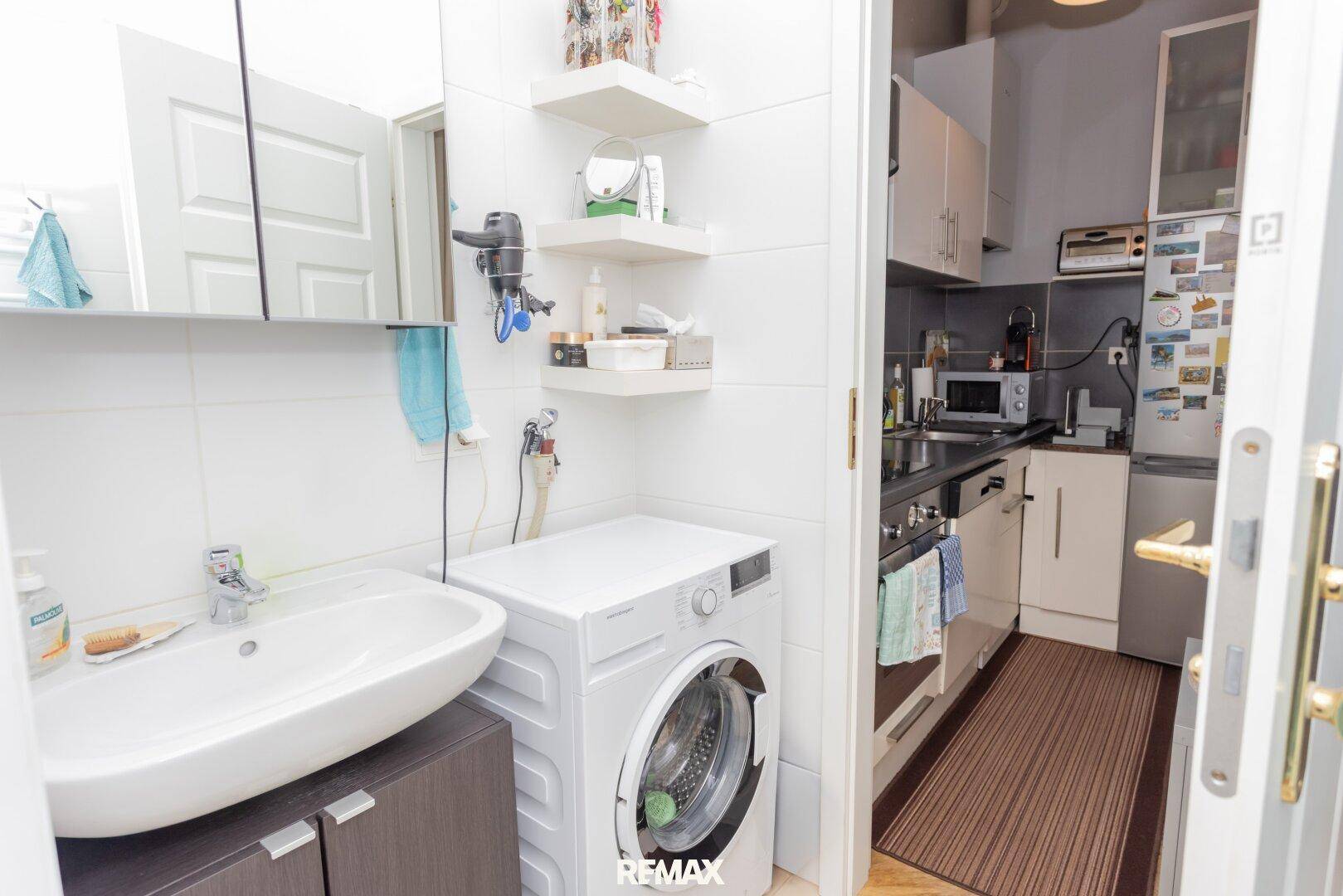 Helles Badezimmer mit Waschbecken, Waschmaschine und Blick in die angrenzende Küche.