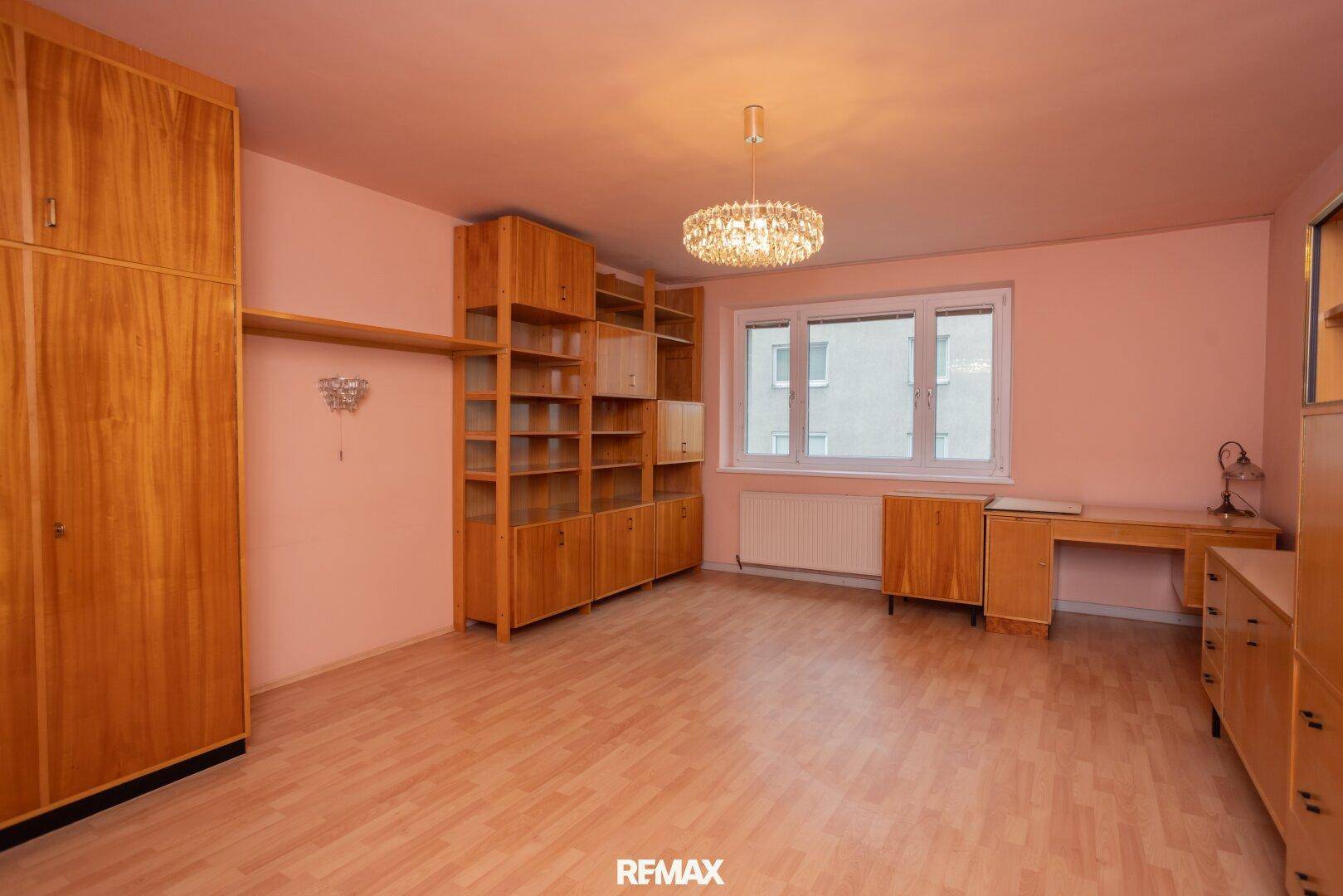 Geräumiges Zimmer mit rosa Wänden, Holzmöbeln und Laminatboden, ausgestattet mit einem großen Fenster.