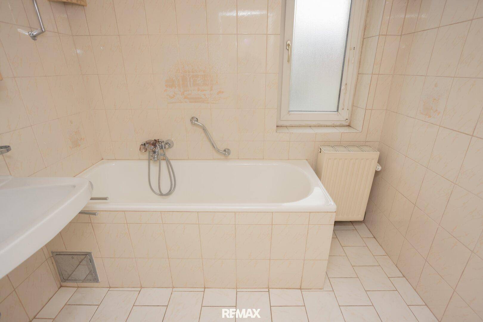 Badezimmer mit Badewanne, beigen Fliesen und einem Fenster, das für natürliche Belüftung sorgt.