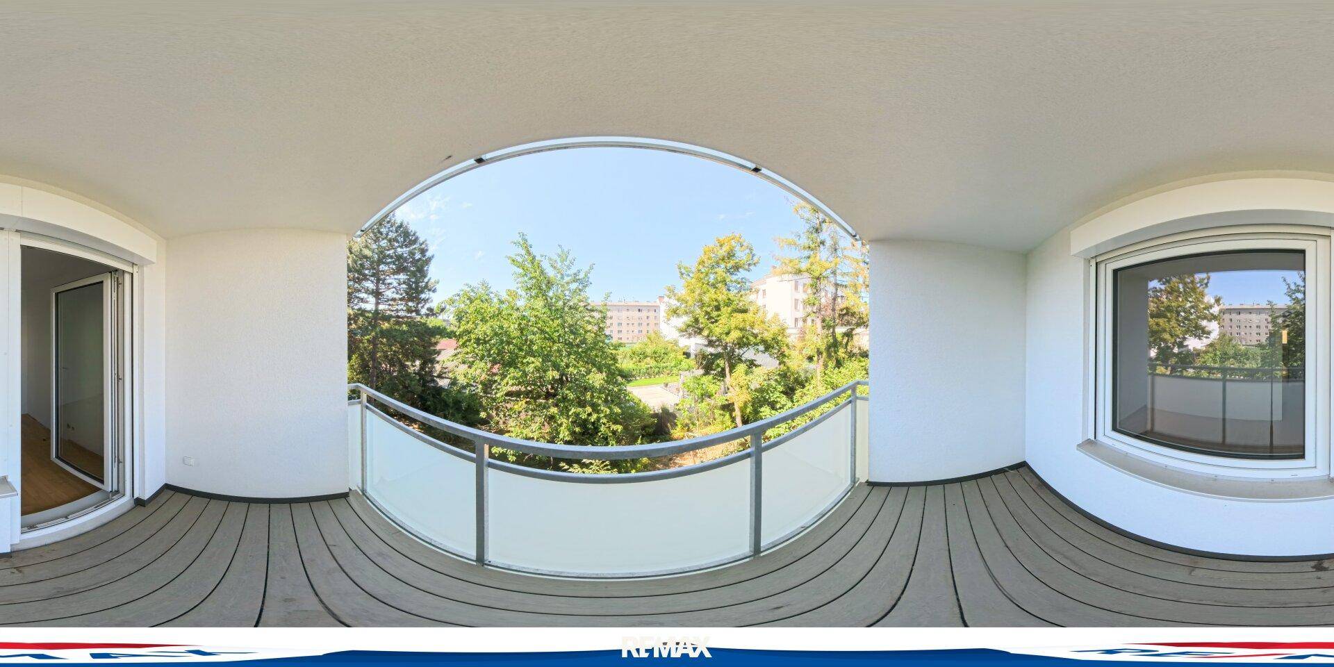 Balkon mit Glasgeländer, Holzboden und Blick ins Grüne sowie auf umliegende Gebäude.