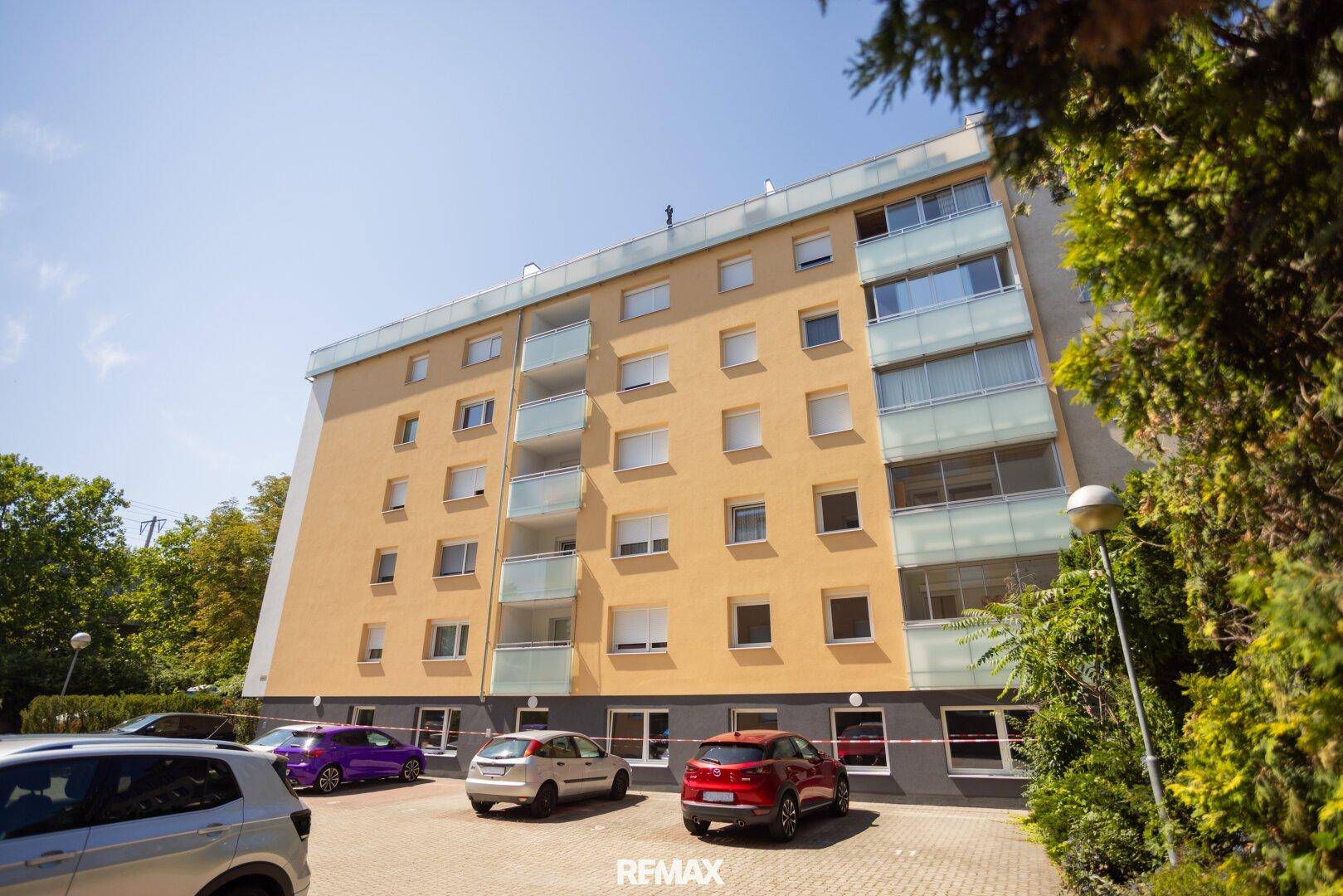 Wohngebäude mit gelber Fassade, vielen Fenstern und Balkonen, umgeben von Bäumen.