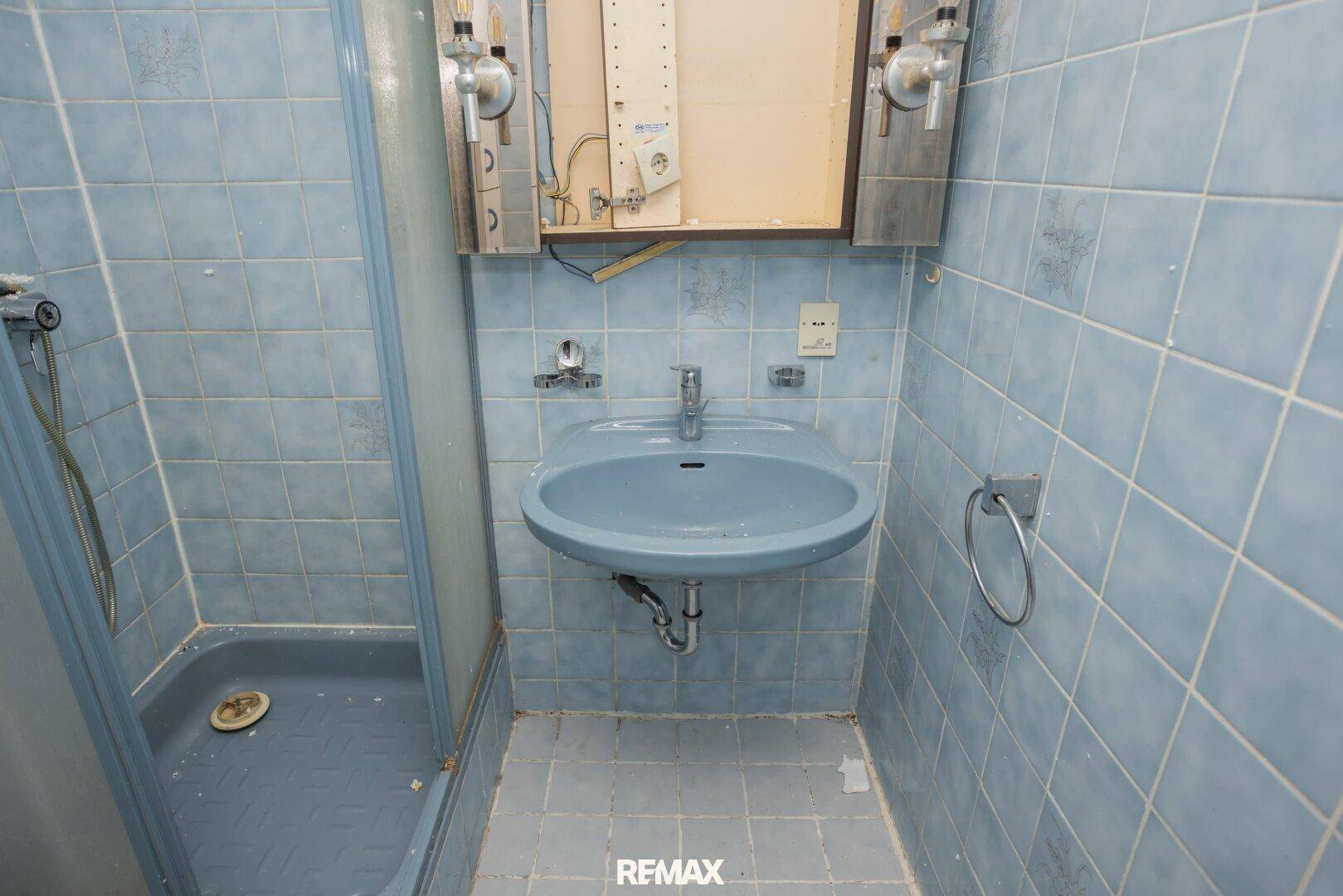 Kleines Badezimmer mit blauen Fliesen, Waschbecken und Duschkabine, renovierungsbedürftig.
