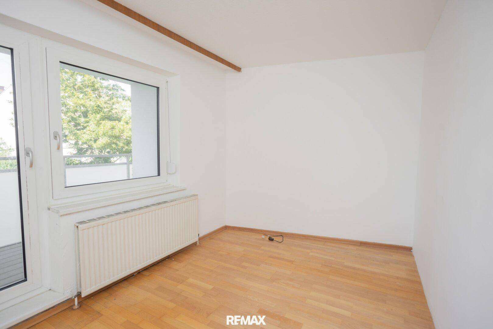 Helles Schlafzimmer mit Holzboden und Fenster mit Blick ins Grüne, unmöbliert.
