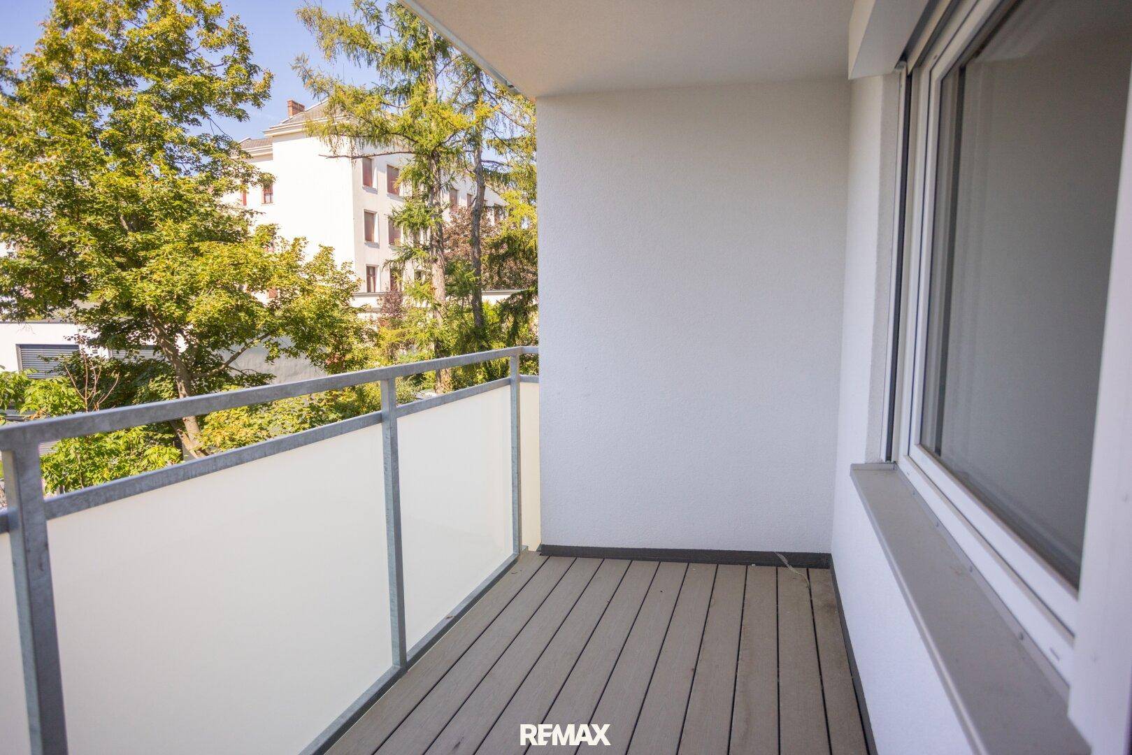 Balkon mit Holzboden und Glasgeländer, bietet einen Blick auf die Umgebung und Bäume.