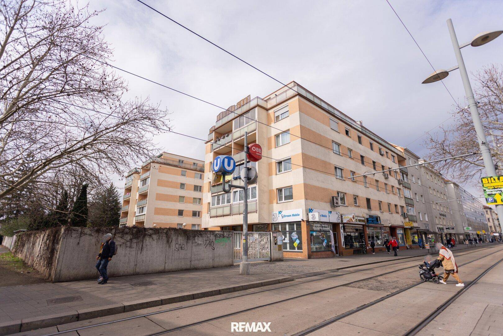 Mehrstöckiges Wohngebäude mit Geschäften im Erdgeschoss und Straßenbahnhaltestelle in urbaner Umgebung.