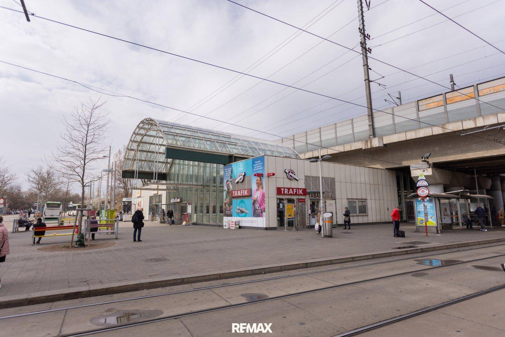 Moderne öffentliche Verkehrsstation mit Glasdach und Geschäften, umgeben von Straßenbahnschienen.