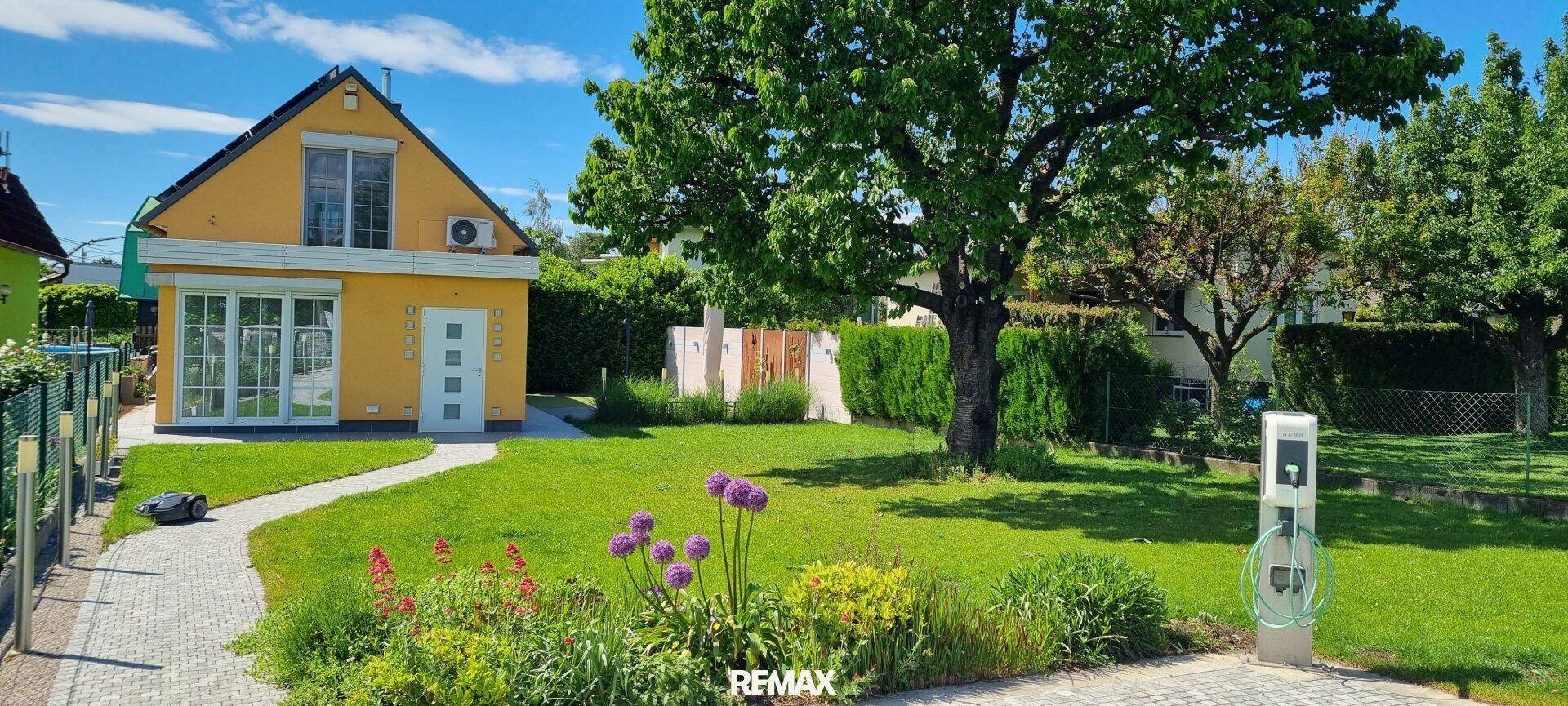 Gelbes Haus mit gepflegtem Garten, großem Baum und Ladestation.