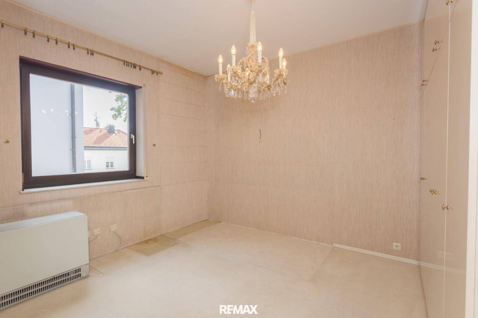 Schlafzimmer mit heller Tapete, Heizkörper und einem eleganten Kronleuchter.