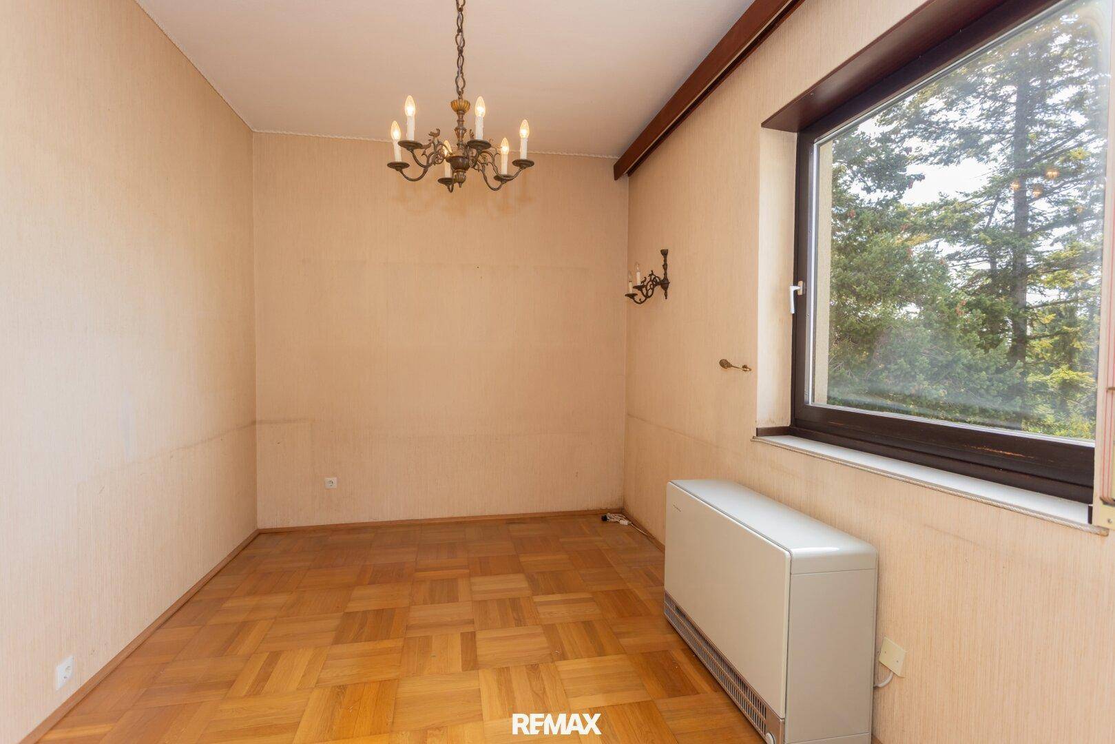 Leeres Zimmer mit Parkettboden, großem Fenster und klassischem Kronleuchter, bereit zur individuellen Gestaltung.