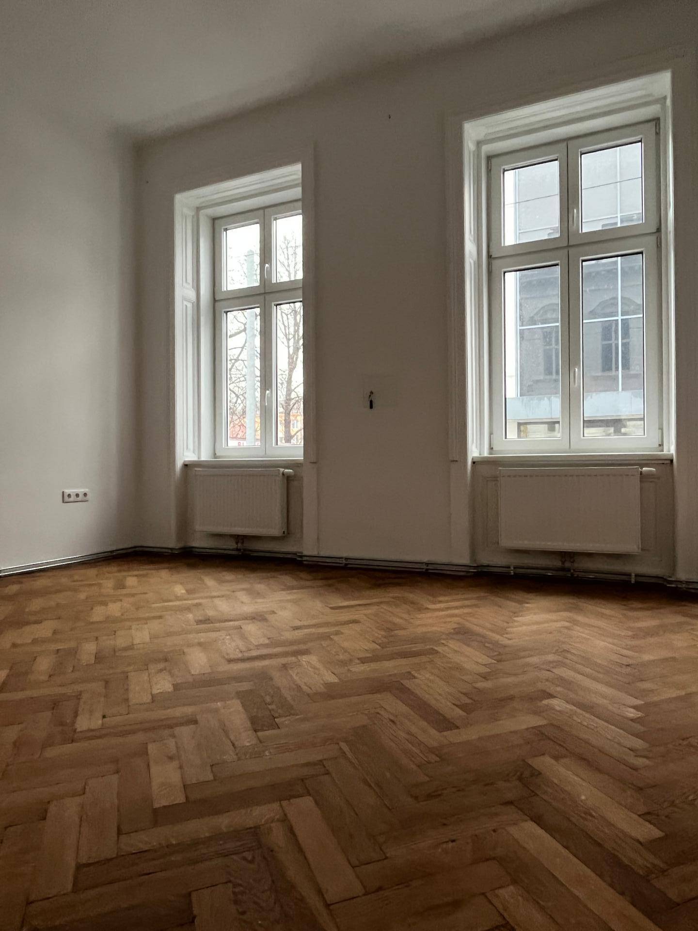 Helles Zimmer mit Fischgrätparkett, hohen Fenstern und Heizkörpern, die für angenehme Wärme sorgen.