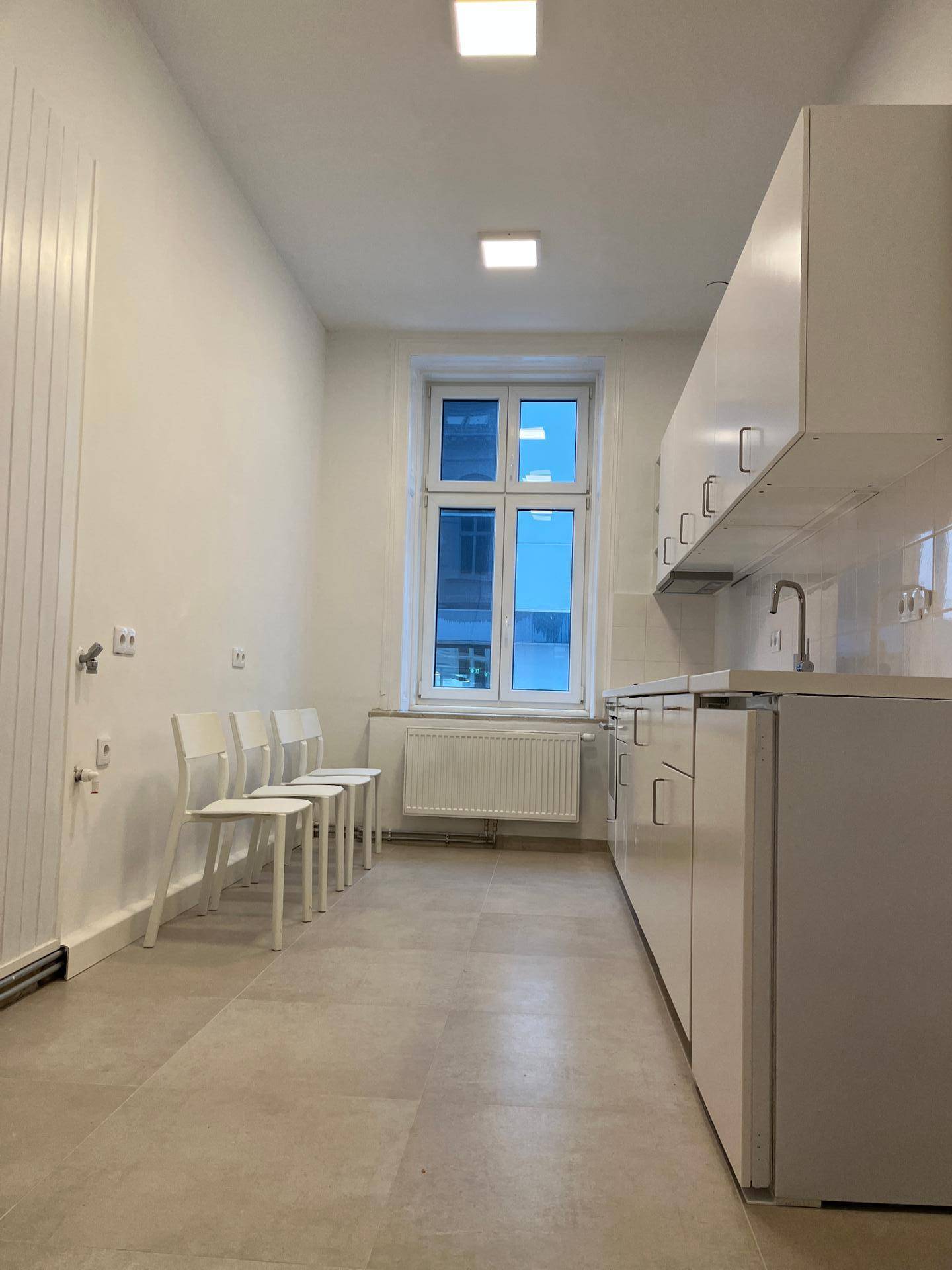 Lange, schmale Küche mit weißen Einbauschränken, Fliesenboden und einem Fenster, das viel Licht hereinlässt.