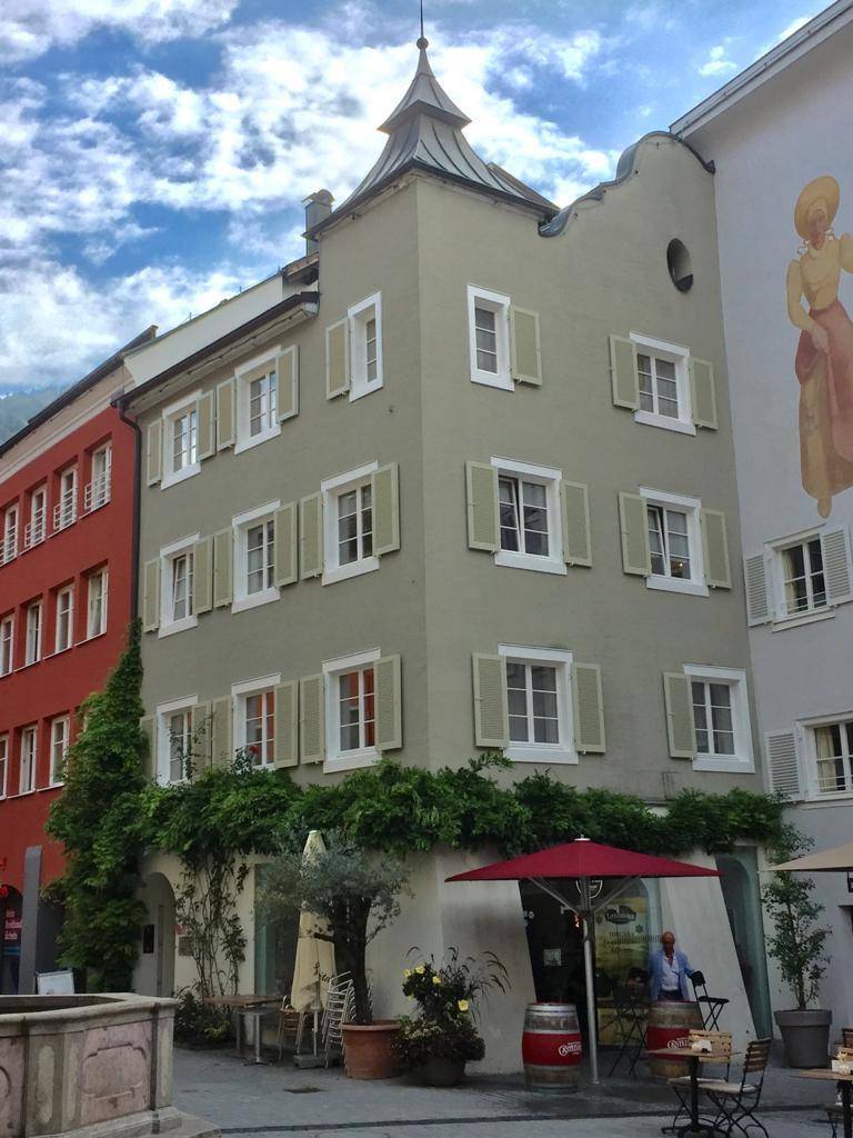 Historisches Gebäude mit traditioneller Architektur, Fensterläden und einem kleinen Außenbereich mit Tischen und Sonnenschirm.
