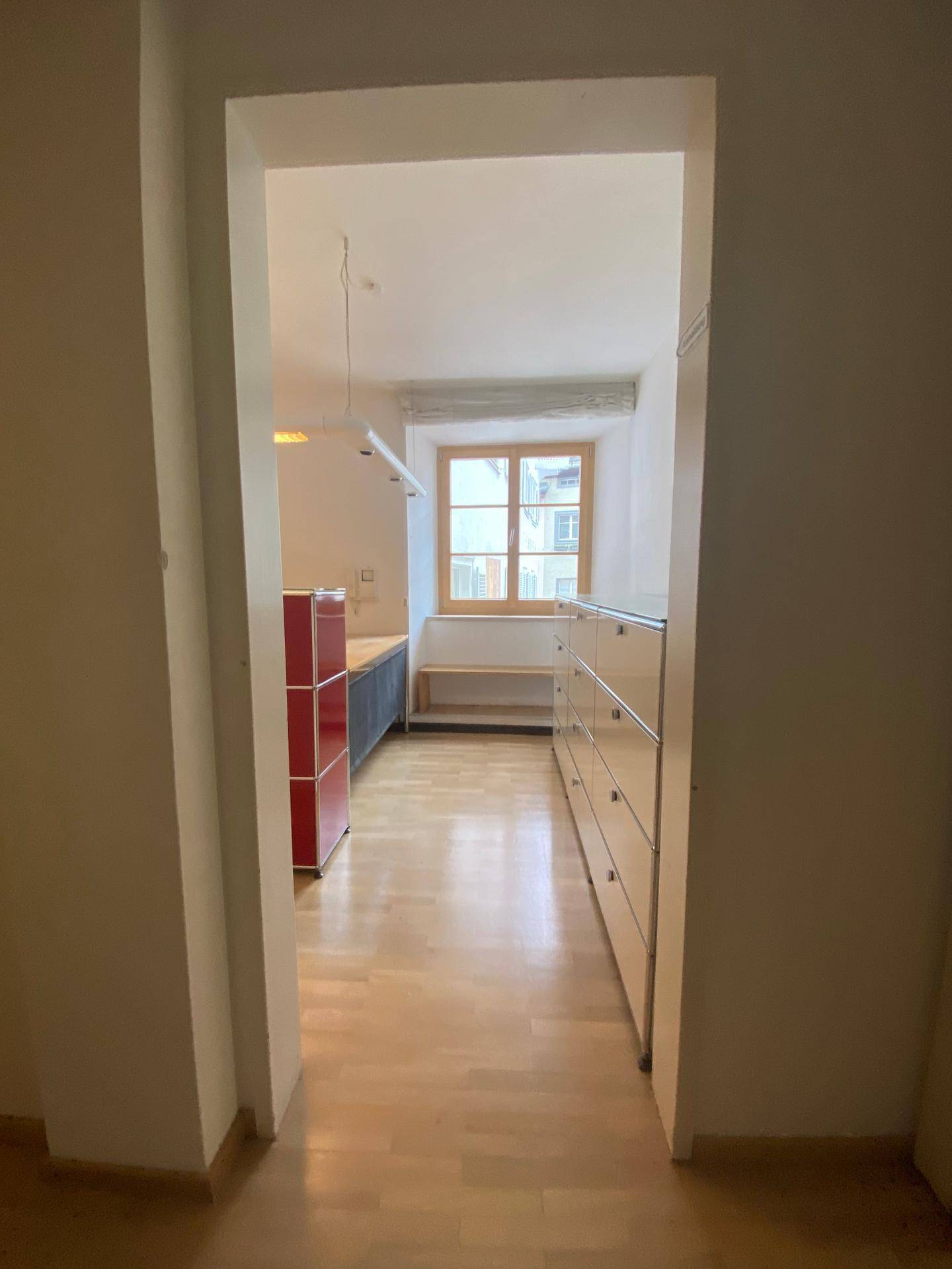 Blick vom Flur in einen hellen Raum mit Holzfenster und Parkettboden, ausgestattet mit modernen Möbeln.