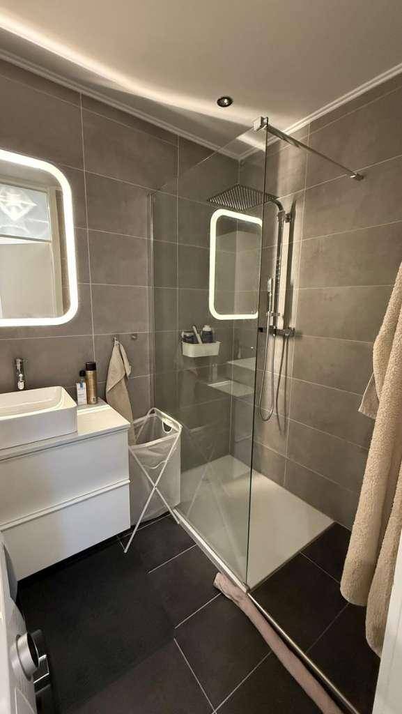 Modernes Badezimmer mit dunklen Fliesen, begehbarer Dusche und beleuchtetem Spiegel.