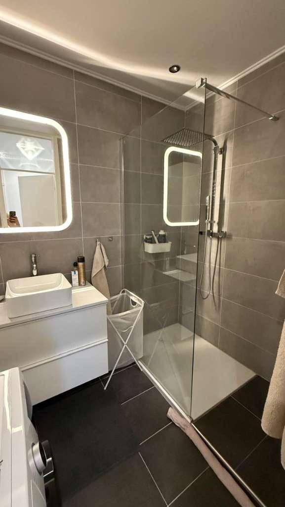 Modernes Badezimmer mit dunklen Fliesen, begehbarer Dusche und beleuchtetem Spiegel.