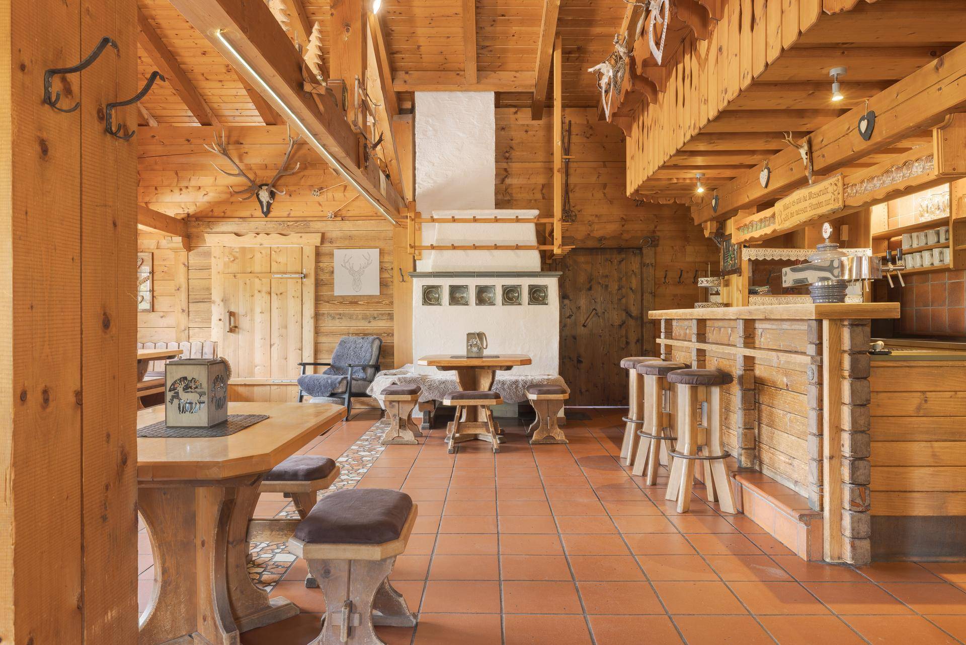 Weitläufiger Speisesaal mit zahlreichen Holztischen und Bänken, einem Kachelofen und einer einladenden Bar.