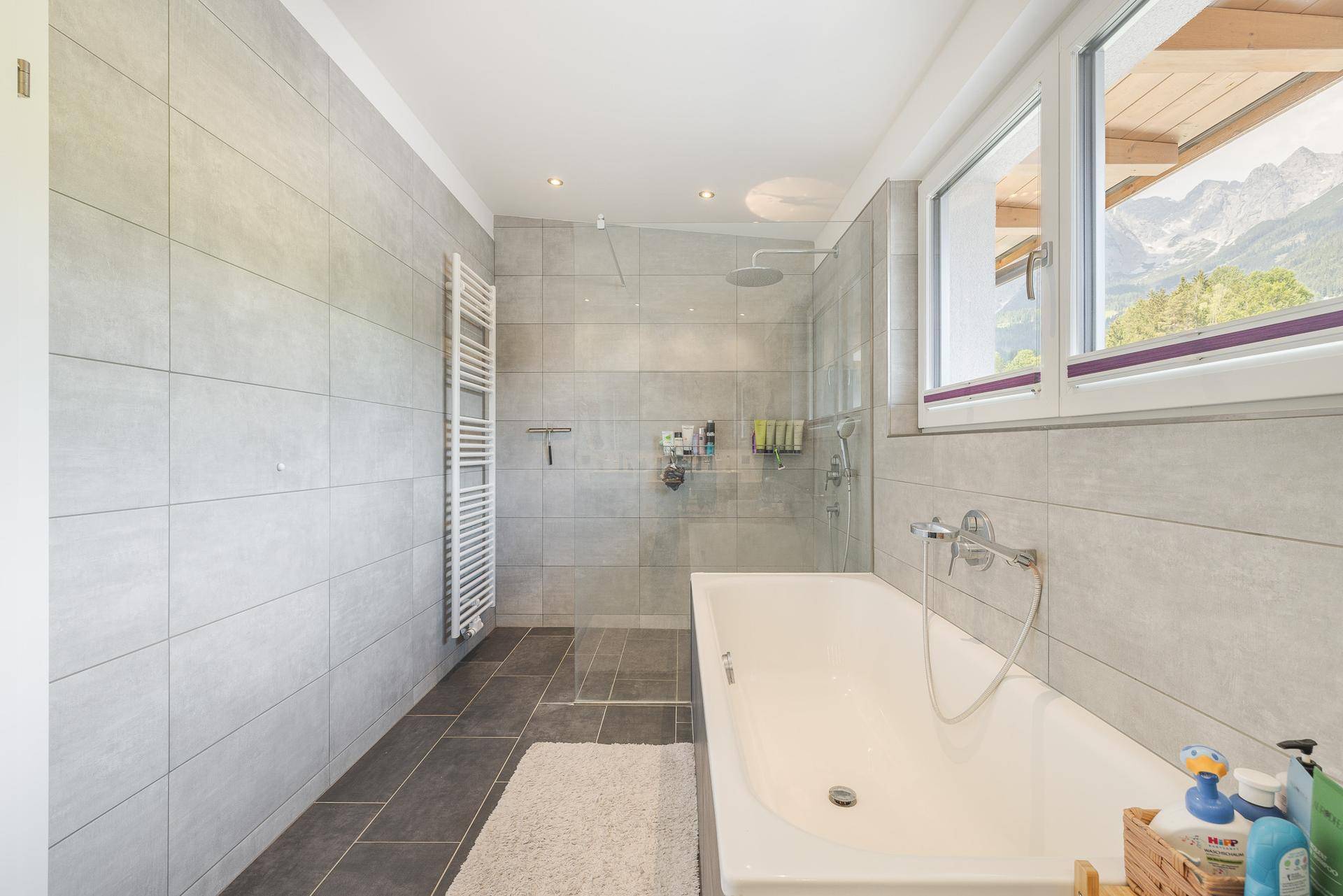 Modernes Badezimmer mit Badewanne, Dusche, Handtuchheizkörper und Bergblick aus dem Fenster.