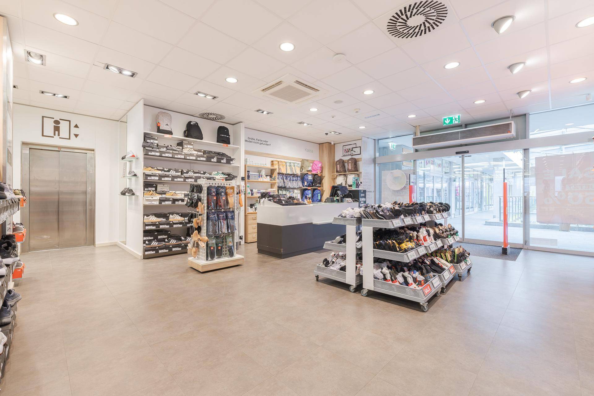 Eingangsbereich eines Schuhgeschäfts mit automatischen Türen, Kasse und Aufzug.