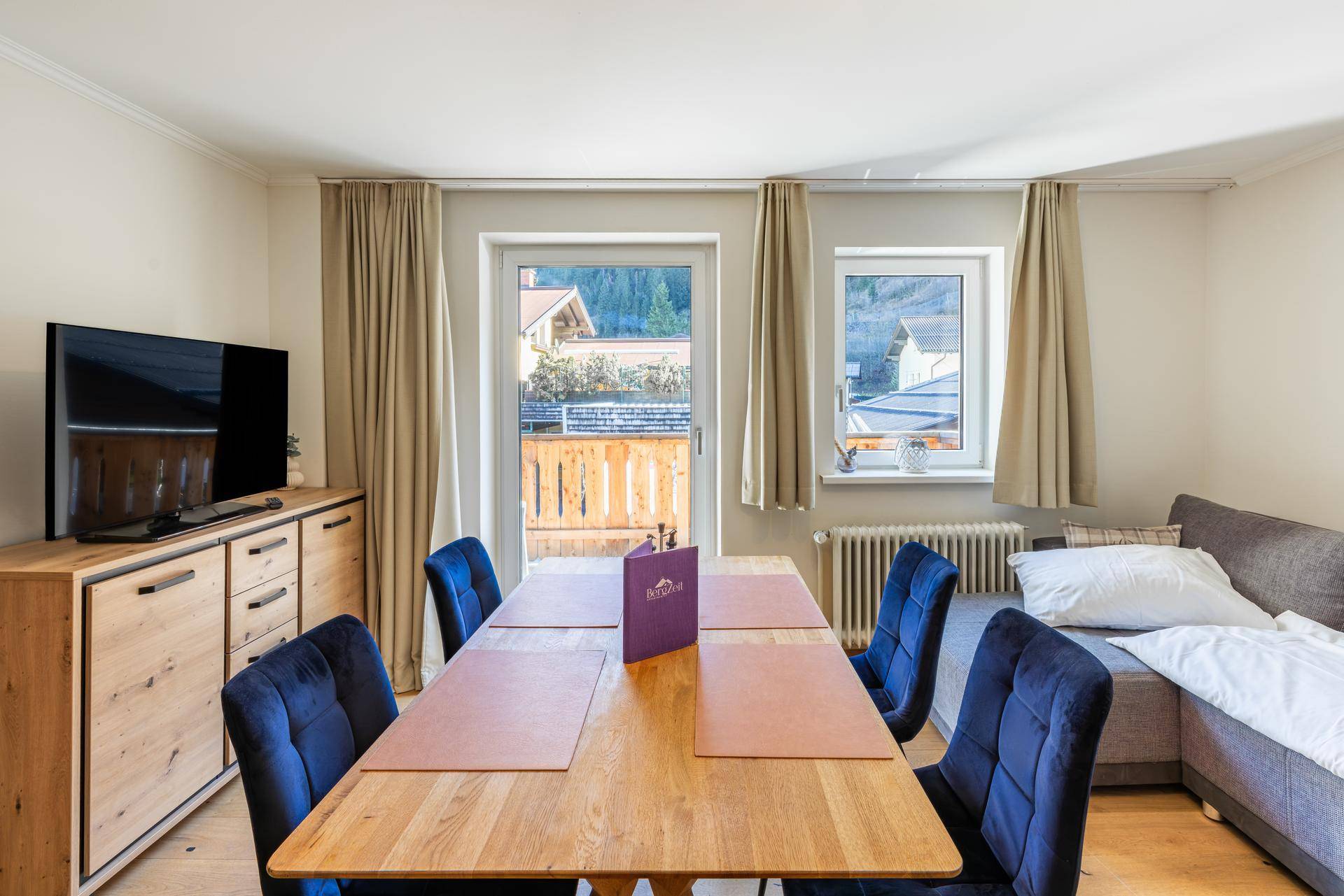 Gemütlicher Wohn- und Essbereich mit Holztisch, blauen Stühlen und Blick auf die Berge.