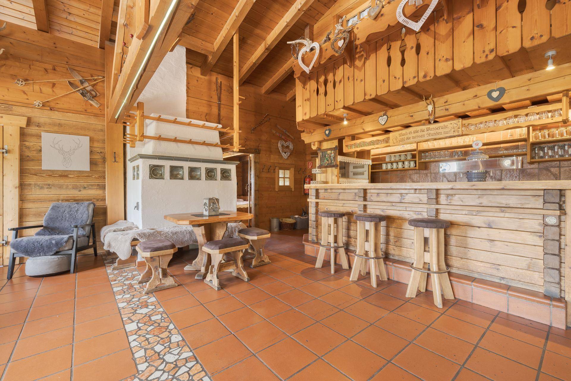 Gemütlicher Innenraum mit traditionellem Kachelofen, rustikaler Holzbar und passenden Hockern, alles in warmem Holz gehalten.