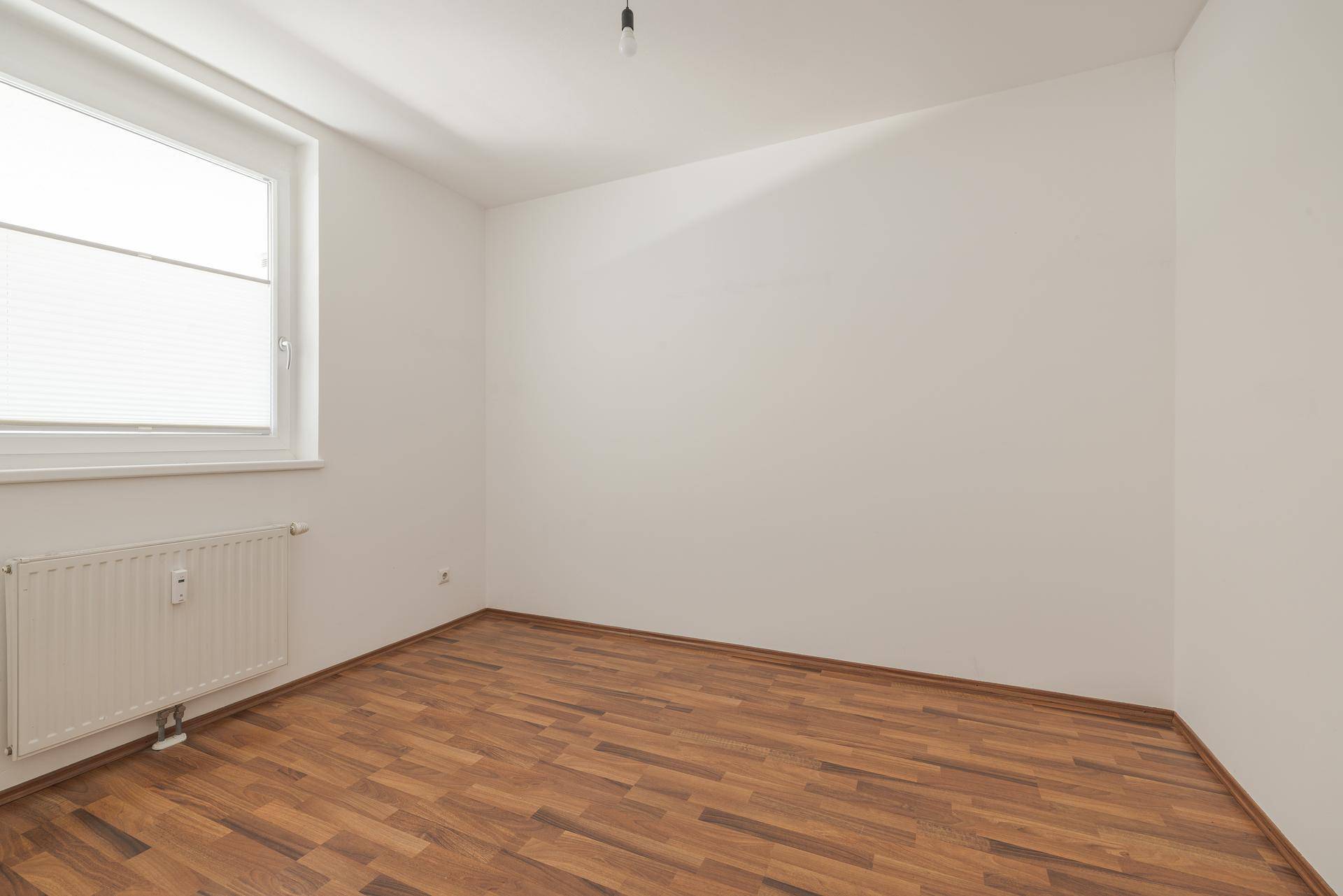 Leeres Schlafzimmer mit Laminatboden, weißen Wänden und einem Fenster mit Jalousien.