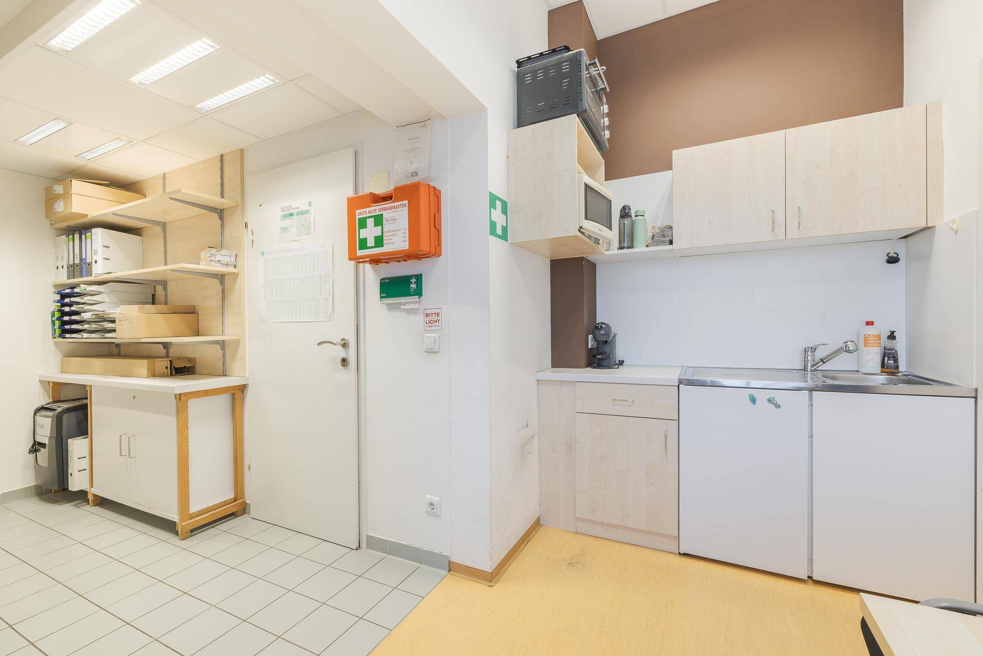 Kleine Mitarbeiterküche mit Spüle, Mikrowelle und Schränken, daneben ein Arbeitsbereich.