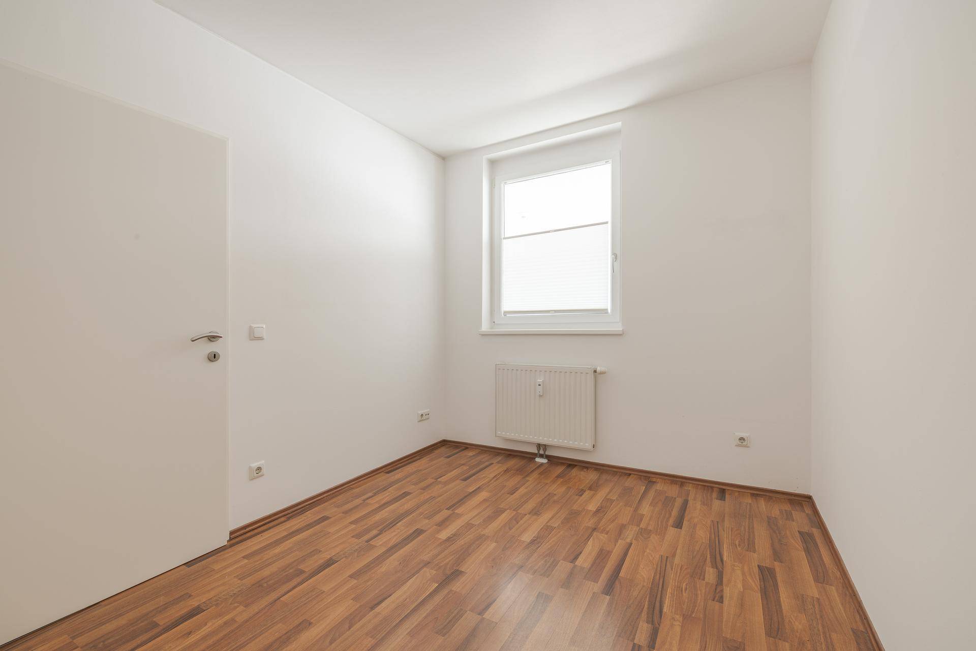 Helles Zimmer mit Laminatboden, weiß gestrichenen Wänden und einem Fenster mit Heizkörper darunter.