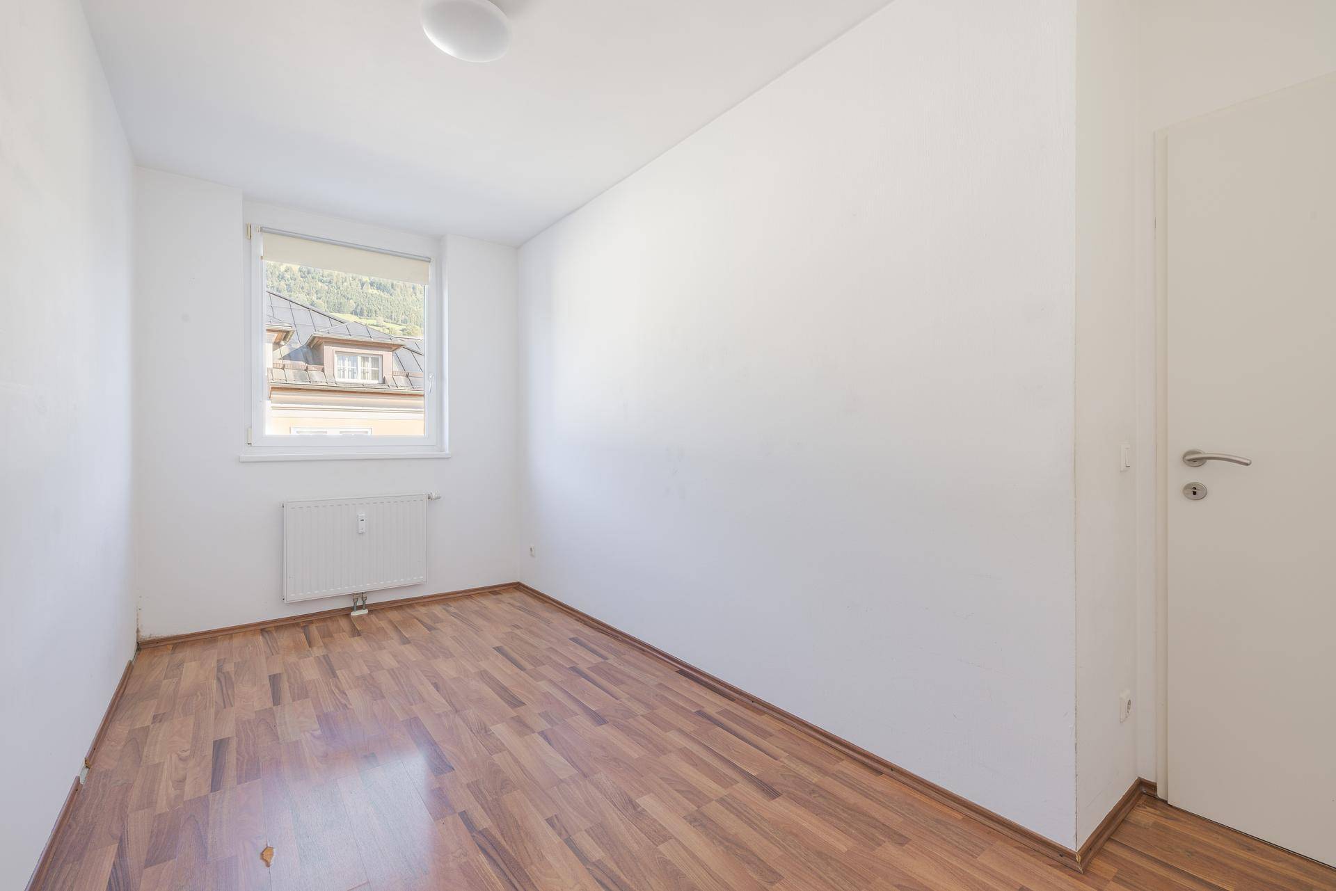 Leeres Zimmer mit Laminatboden, weißen Wänden und einem Fenster mit Blick auf die Umgebung.