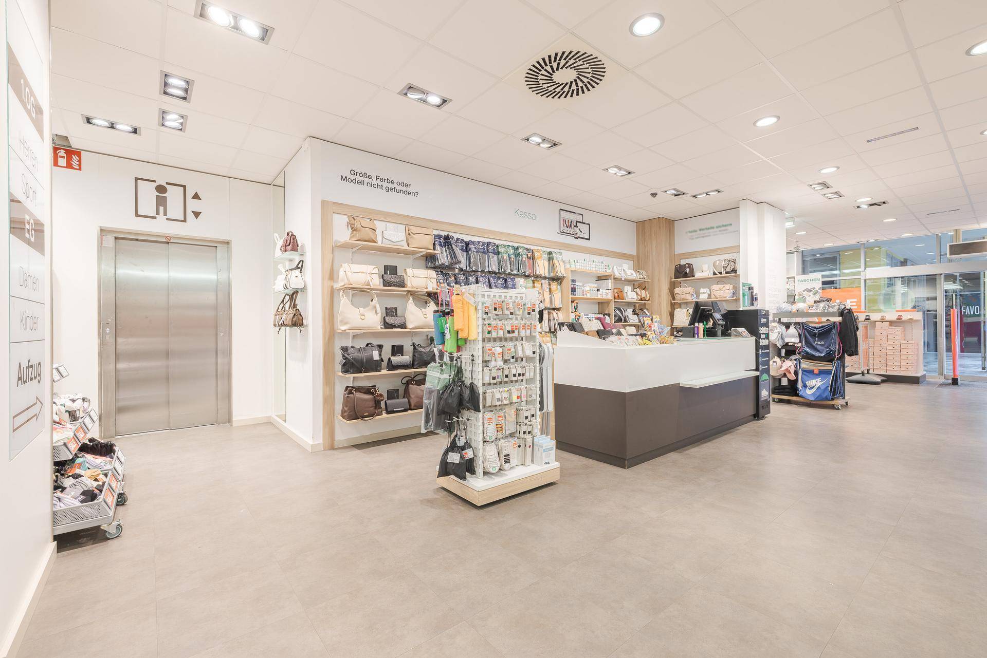 Eingangsbereich eines Schuhgeschäfts mit Kasse, Aufzug und Auslagen für Accessoires.