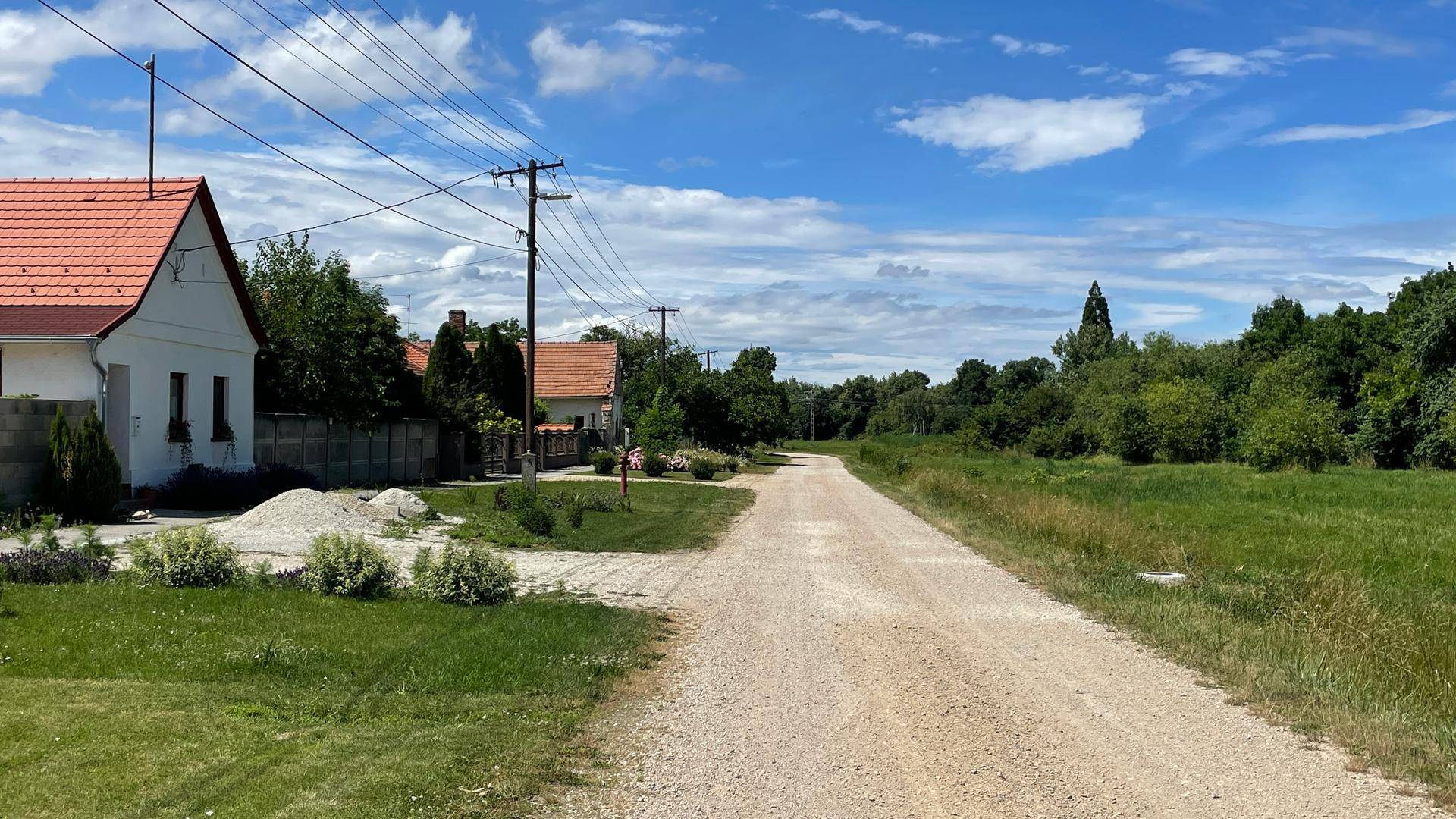 Ländlicher Feldweg führt an traditionellen Häusern vorbei, umgeben von grünen Wiesen und Bäumen.