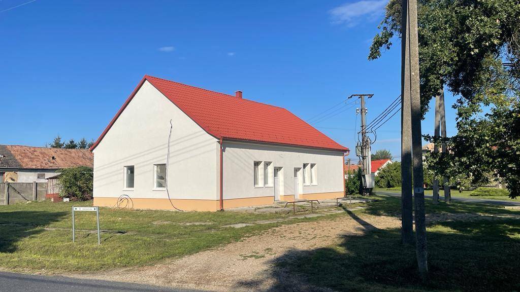 Das Haus von der Straße aus gesehen, mit rotem Dach, weißer Fassade und umgebender Natur.