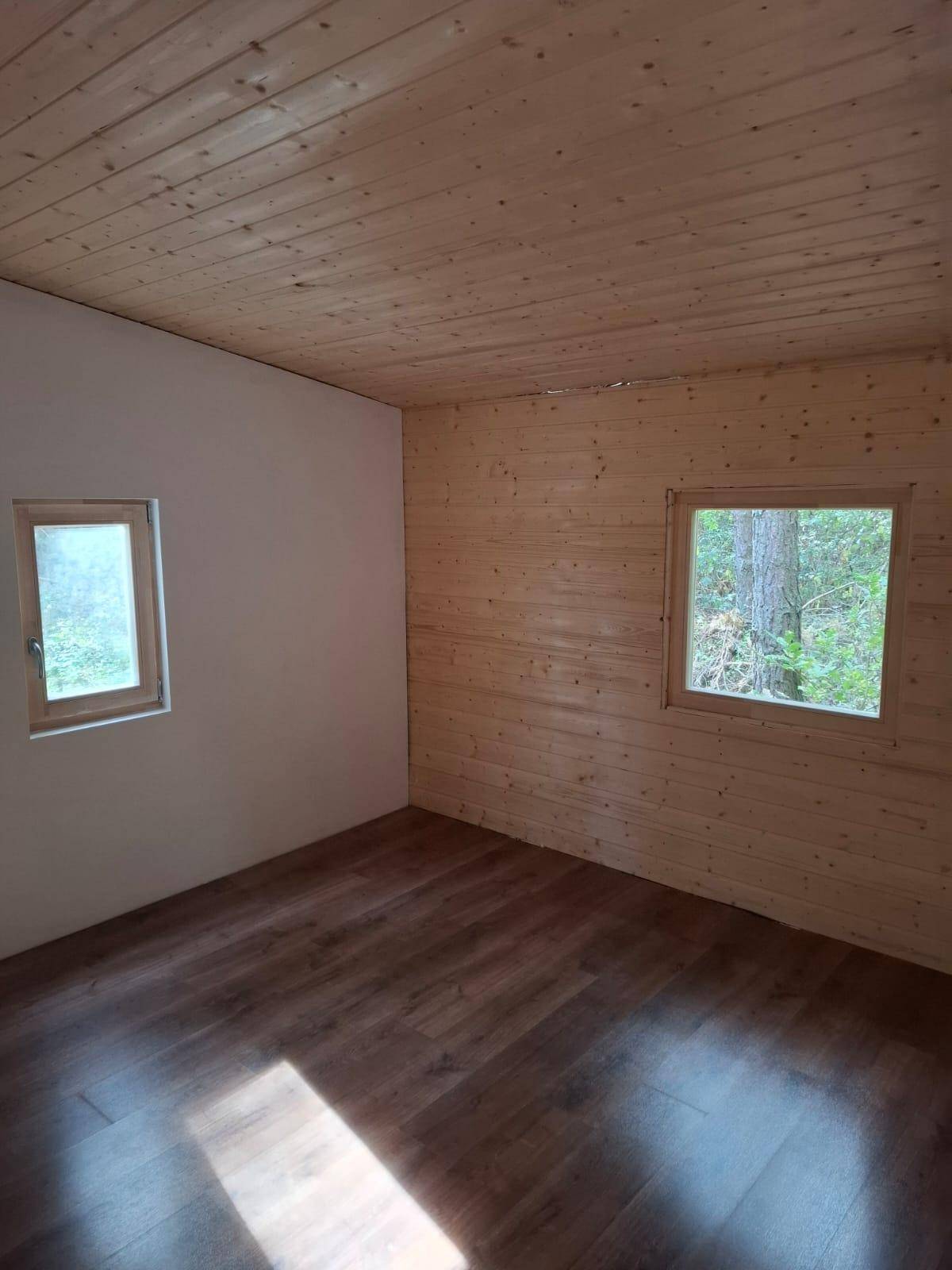 Heller Innenraum mit Holzwänden und -decke, Laminatboden und Blick ins Grüne durch die Fenster.