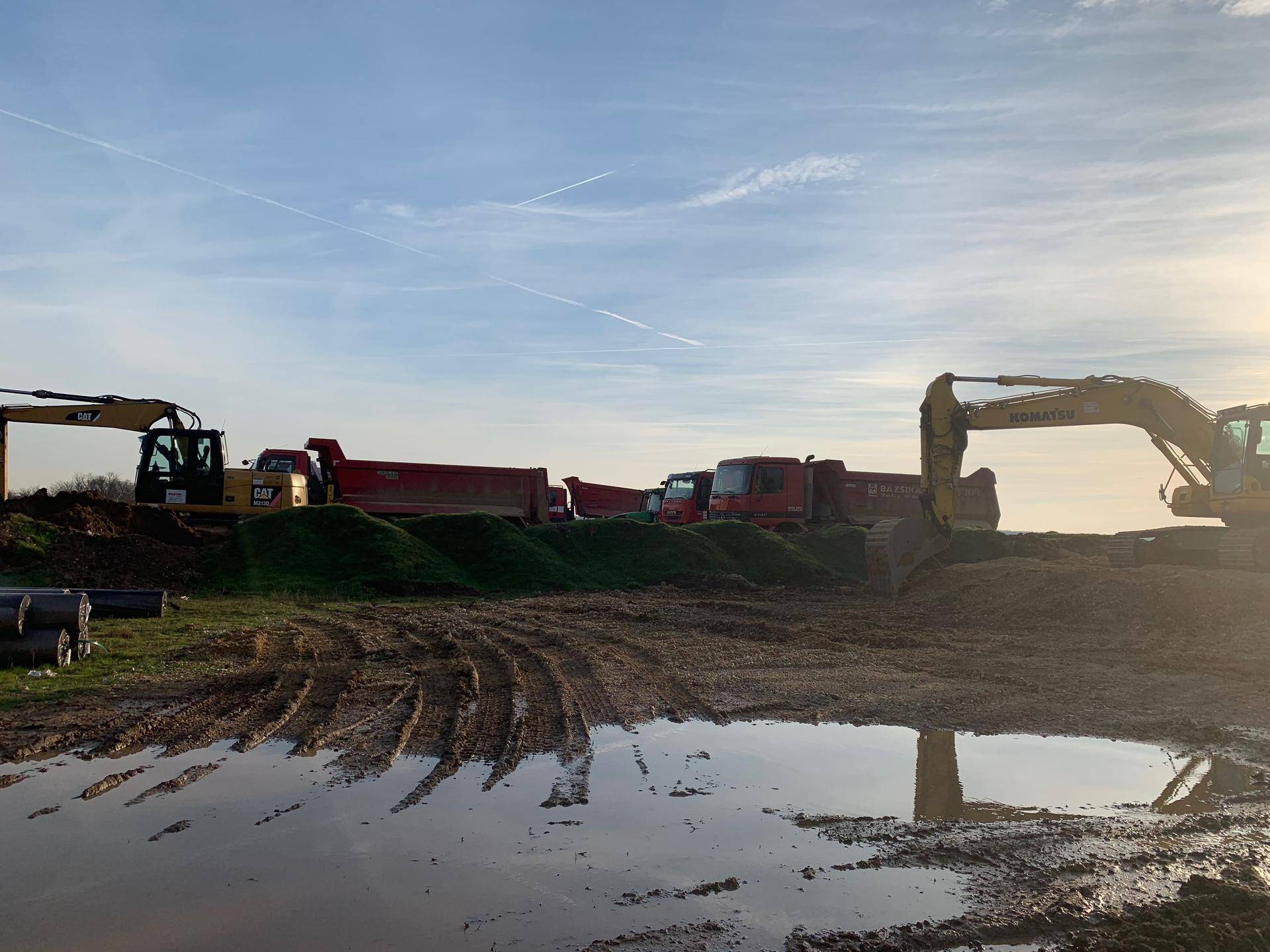 Baustelle bei Sonnenuntergang mit Baggern und Lastwagen auf schlammigem Gelände und Pfützen.