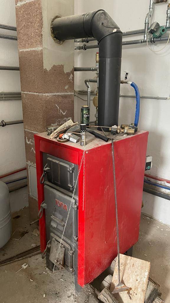 Heizungsanlage mit einem roten Festbrennstoffkessel und sichtbaren Rohren in einem Technikraum.
