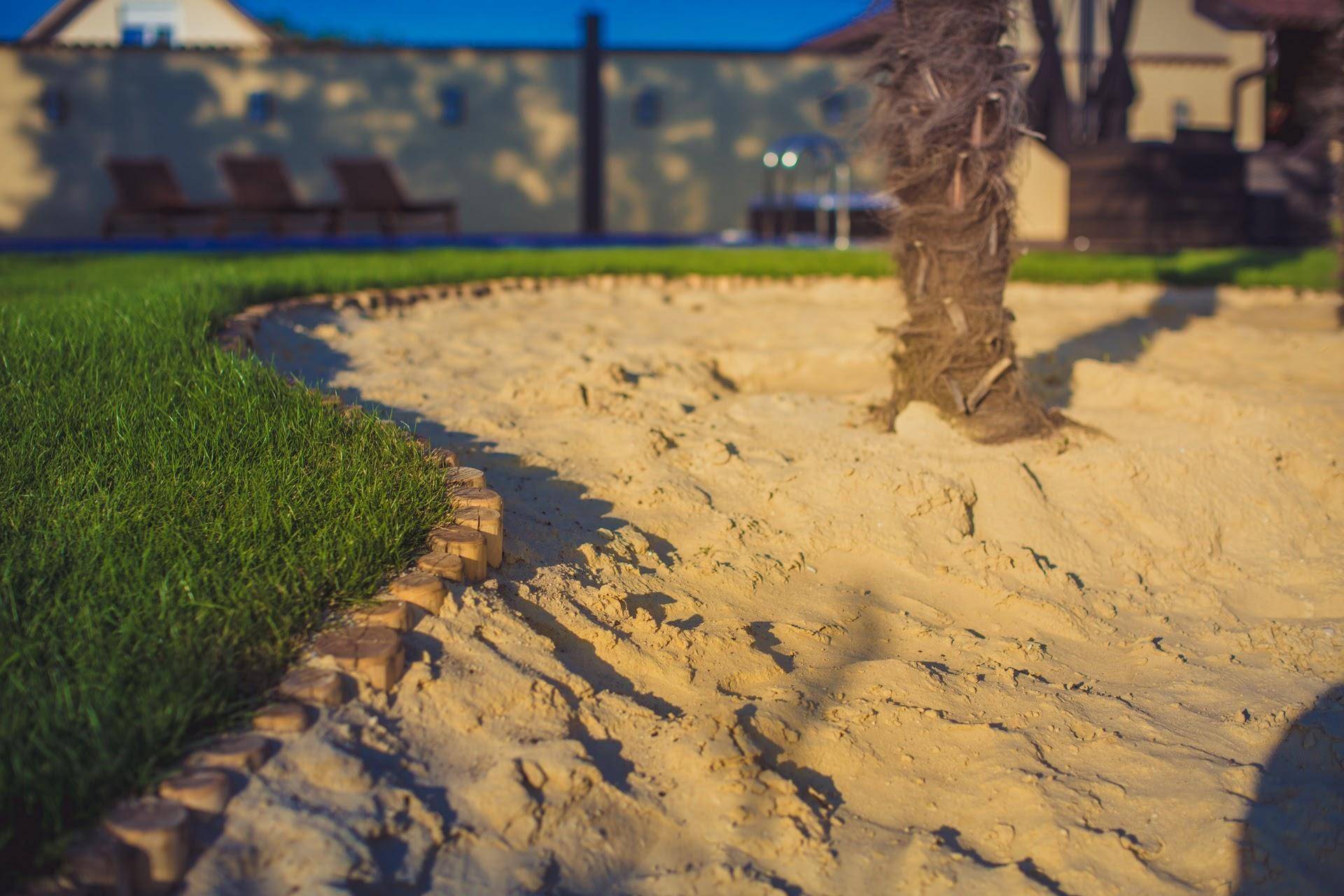 Detailaufnahme eines Sandbereichs mit Palmen und Rasen im Garten, bei warmem Abendlicht.