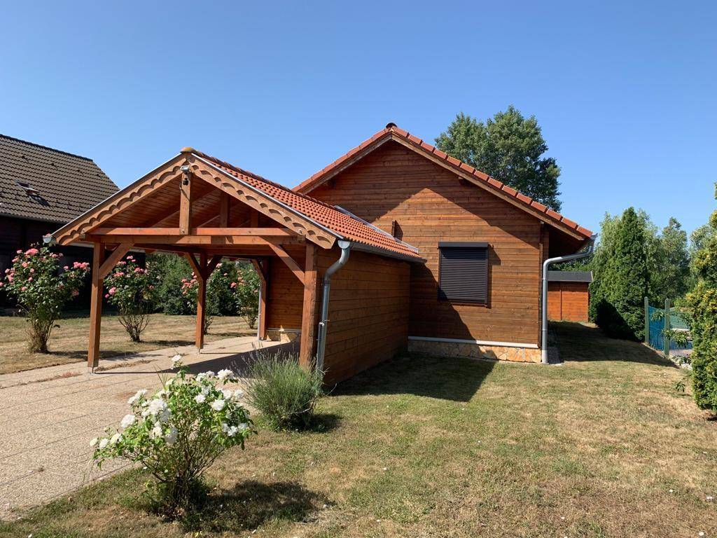 Holzhaus mit Carport und gepflegtem Garten unter strahlend blauem Himmel, inklusive Rosenbeeten.