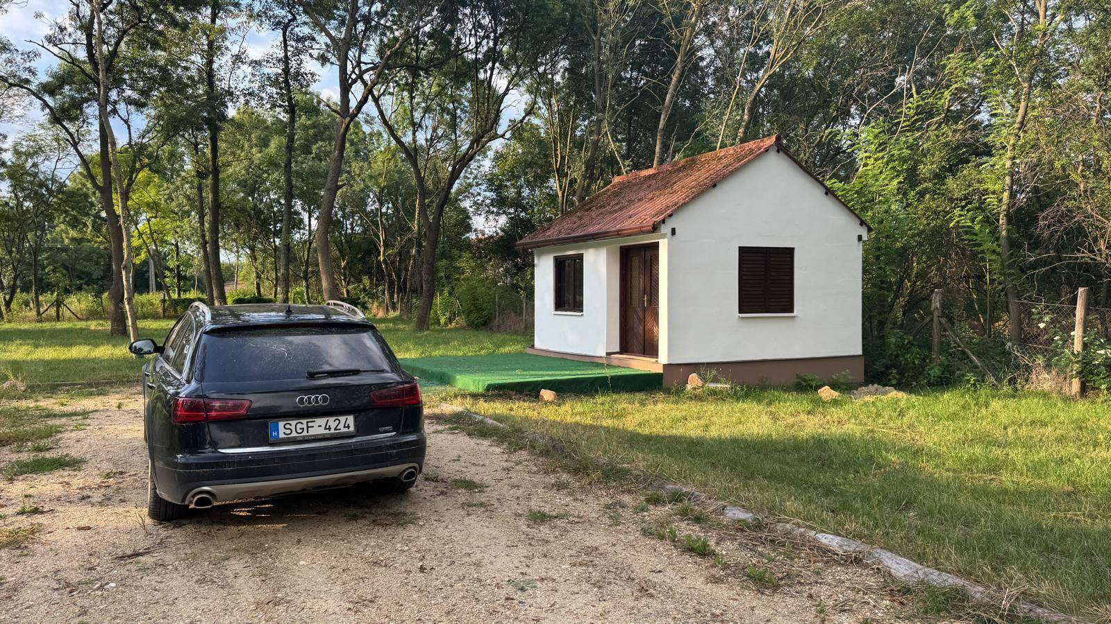 Kleines Haus mit rotem Dach, umgeben von Bäumen, mit einem geparkten Auto auf dem Schotterweg.