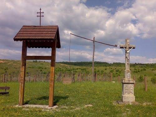 Ländliche Szene mit einem Holzkreuz und einem kleinen Unterstand in einer grünen Landschaft.
